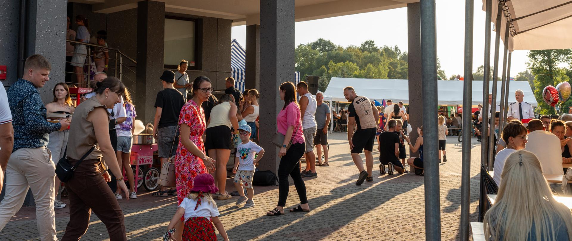 Na zdjęciu uczestnicy pikniku przy remizie OSP Przytkowice. Przed budynkiem, na którym widoczny jest czerwony napis „OSP PRZYTKOWICE”, zgromadzili się dorośli i dzieci. W tle znajdują się namioty, stoiska oraz osoby uczestniczące w wydarzeniu. Fotografia została wykonana w słoneczne, letnie popołudnie.