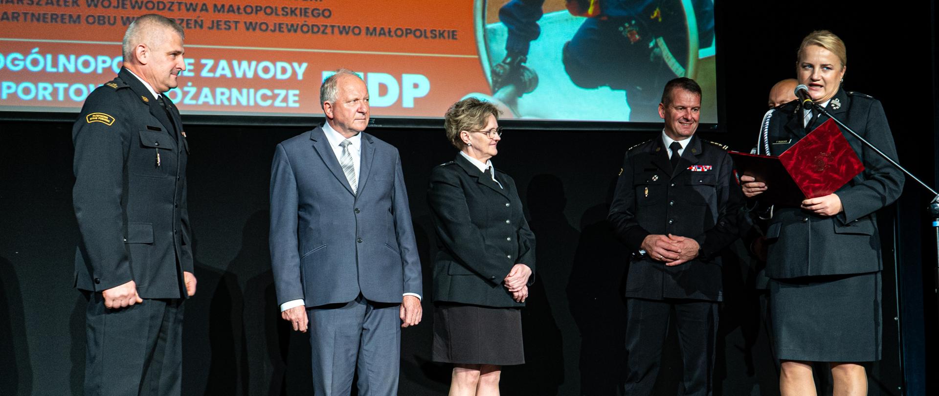 Na scenie sali widowiskowej stoją przedstawiciele gminy i strażaków, za nimi fragment ekran z napisem Ogólnopolskie zawody sportowo-pożarnicze MDP