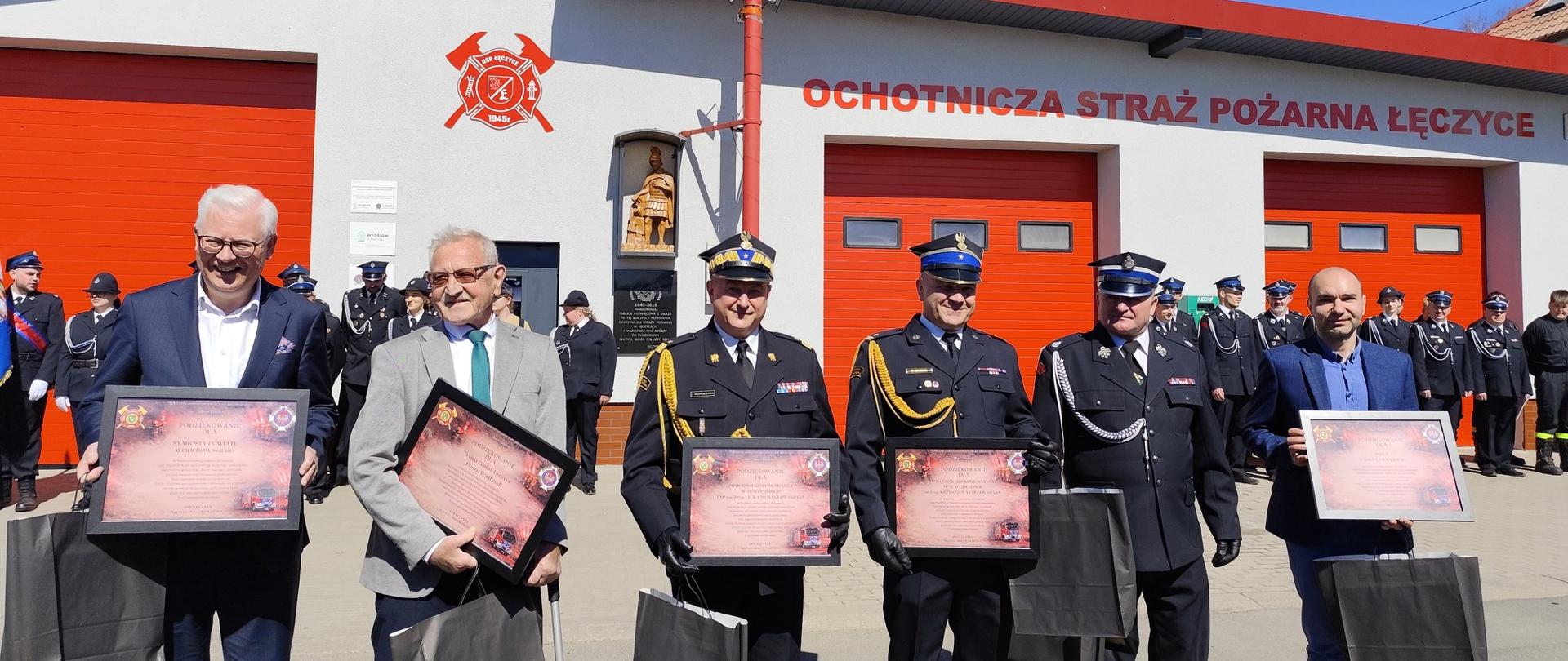 Wójt, zastępca wójta, wicestarosta wejherowski oraz przedstawiciele straży pożarnej pozujący z podziękowaniami w ramkach na tle remizy strażackiej