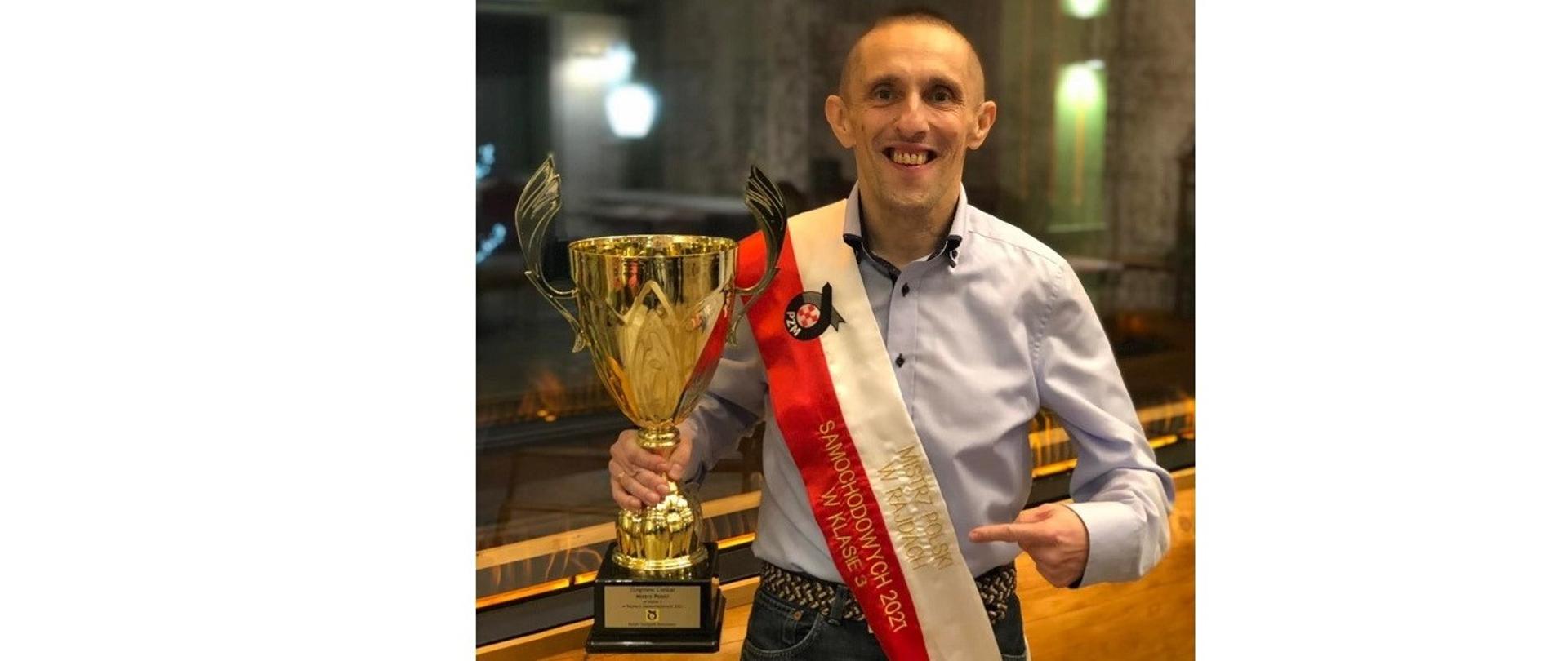 Zbigniew Cieślar z pucharem za zdobycie tytułu Mistrza Polski w Rajdach Samochodowych w Klasie 3, który uzyskał w roku 2021 jako pilot w załodze z kierowcą Łukaszem Byśkiniewiczem. Przepasany biało-czerwoną szarfą Mistrzów Polski mężczyzna trzyma w prawym ręku puchar i wskazuje na niego palcem lewej ręki. Fot. Materiały prasowe 