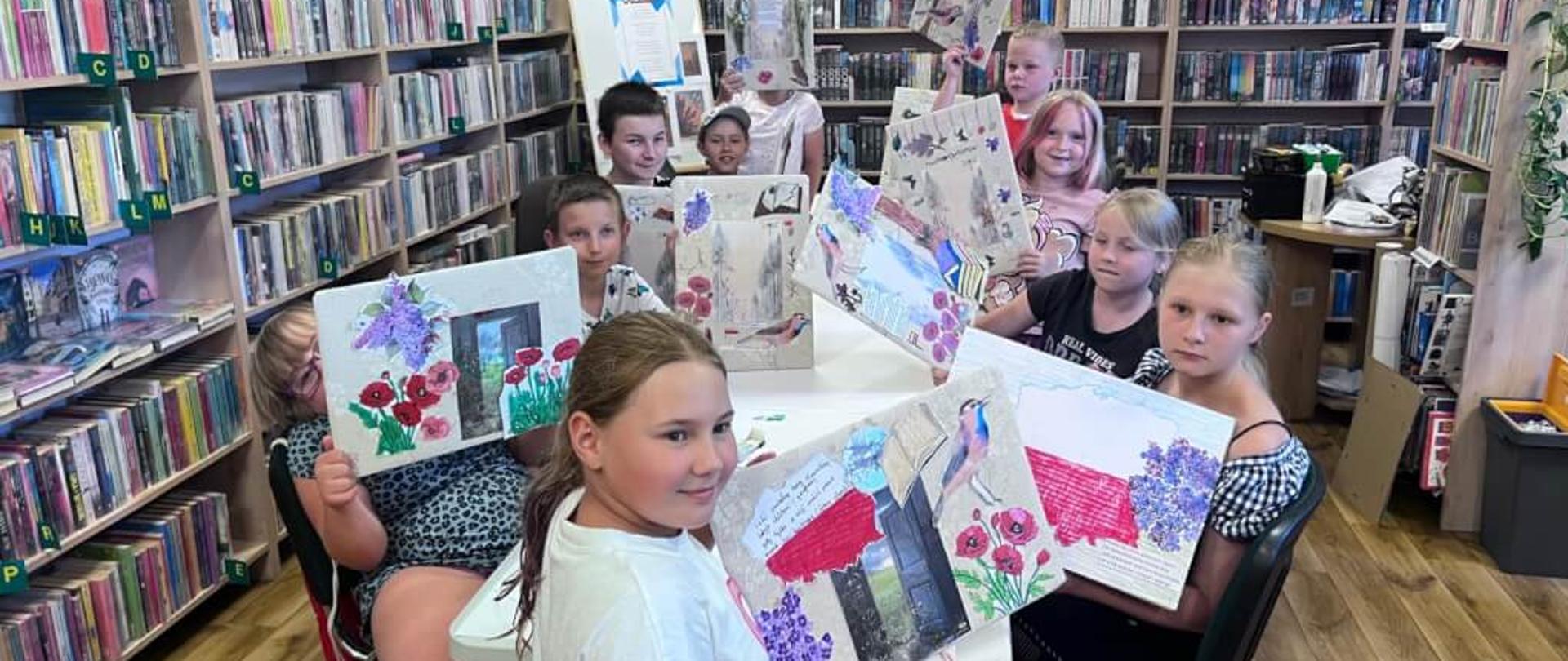 Dzieci w bibliotece siedzą przy dużym stole i pokazują swoje prace plastyczne.