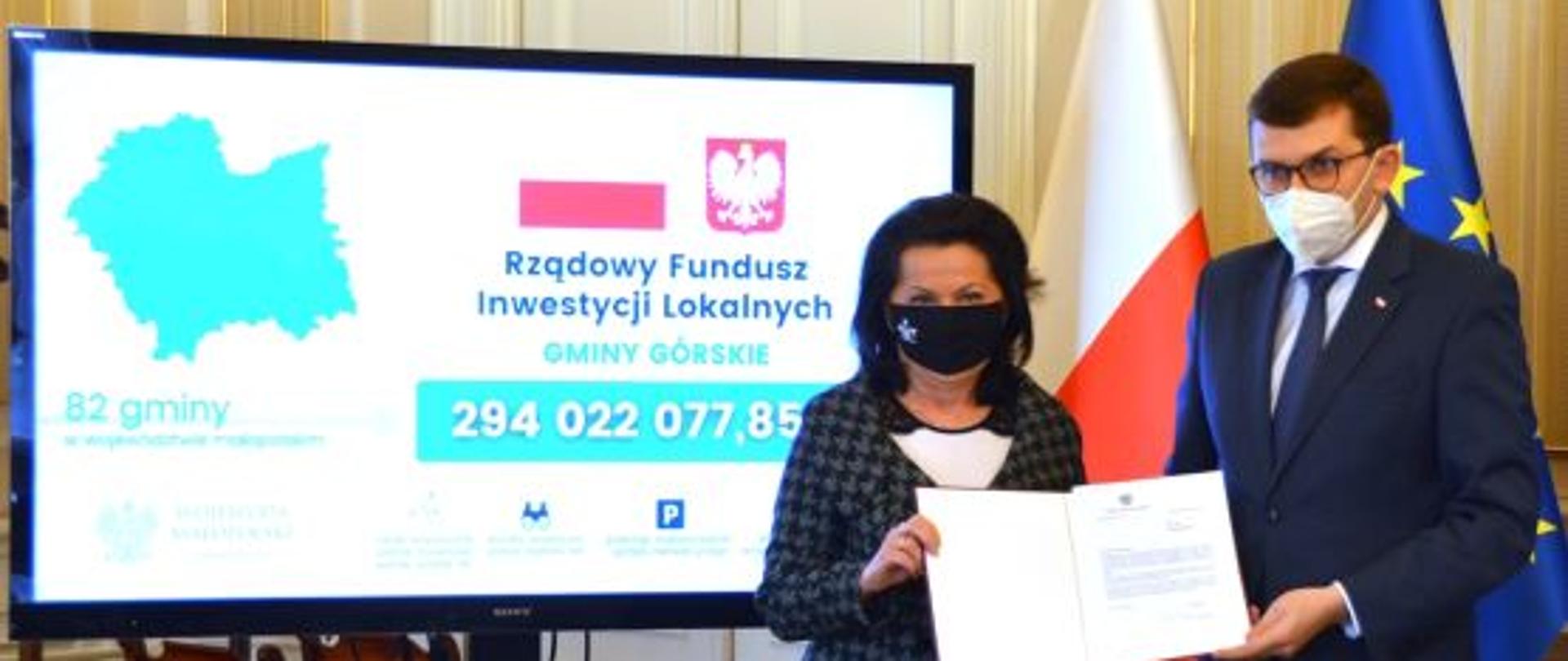 Zdjęcie wykonano w Małopolskim Urzędzie Wojewódzkim w Krakowie wojewoda małopolski Łukasz Kmita wraz z posłanką Anną Paluch prezentują wyniki wyki naboru. Tuż za nimi flaga polski oraz uni europejskiej . Z lewej strony telewizor na którym zostaje wyświetlona kwota dofinansowania wraz z informacjami dotyczącymi Rządowego funduszu inwestycji lokalnych.