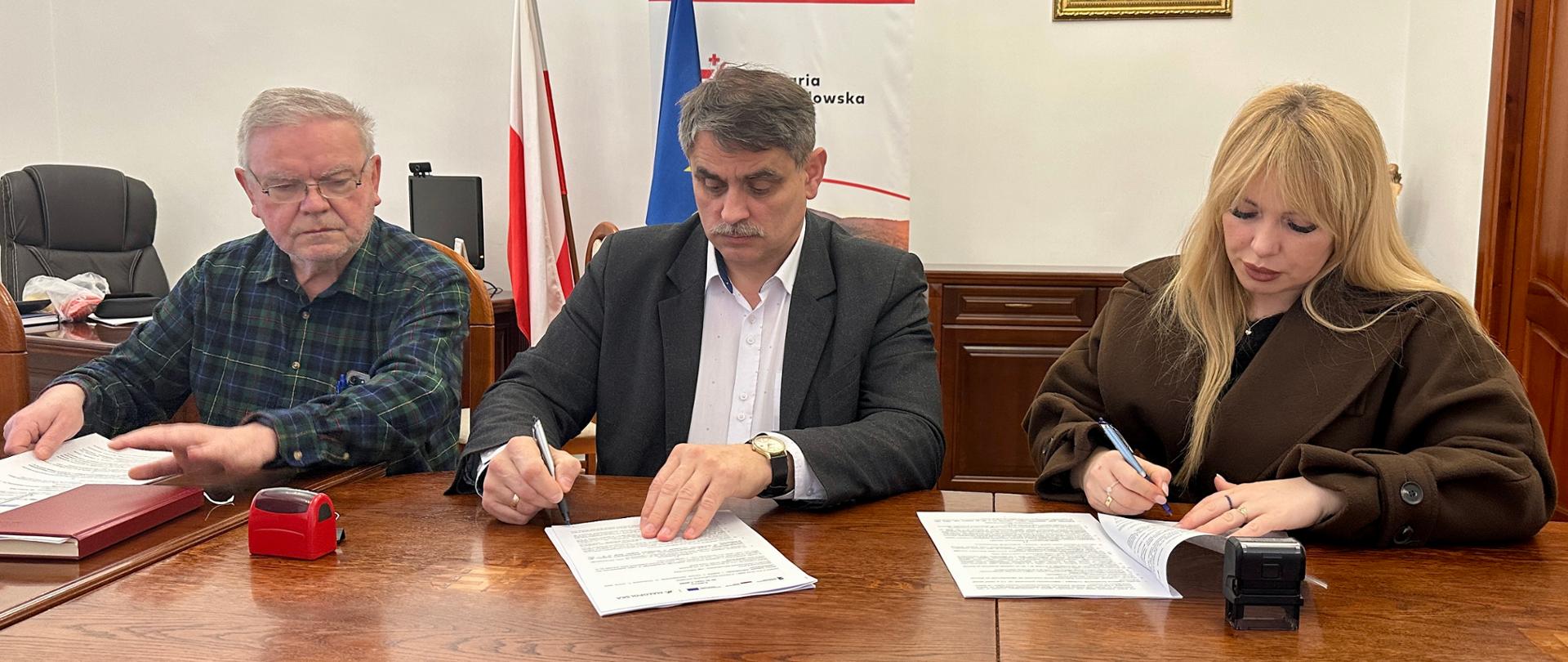 Trzy osoby siedzą przy drewnianym stole w urzędzie i podpisują dokumenty. W centralnej części mężczyzna w garniturze składa podpis, obok niego po lewej starszy mężczyzna przegląda dokumenty, a po prawej kobieta również podpisuje umowę. W tle widoczne są flagi Polski i Unii Europejskiej oraz godło państwowe na ścianie.