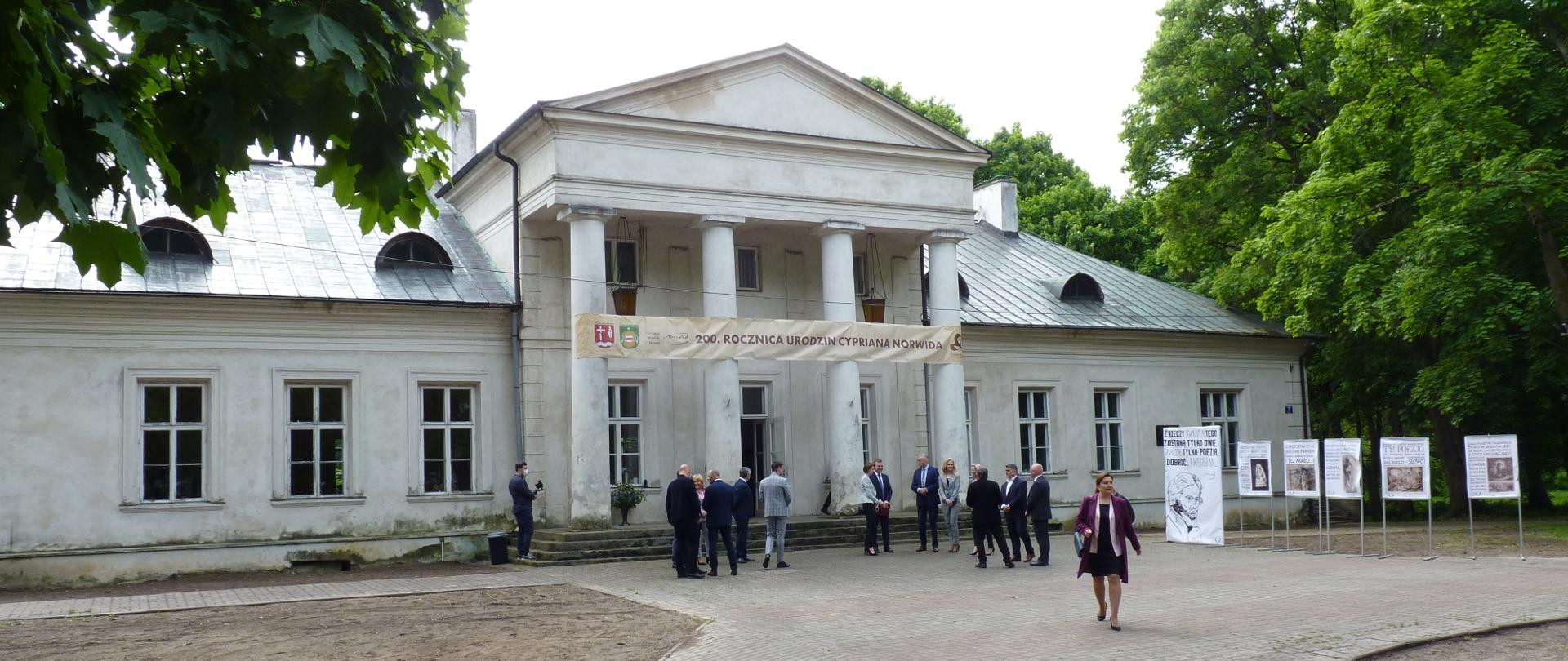 Biały pałac z czterema wysokimi kolumnami. Przed pałacem stoją ludzie