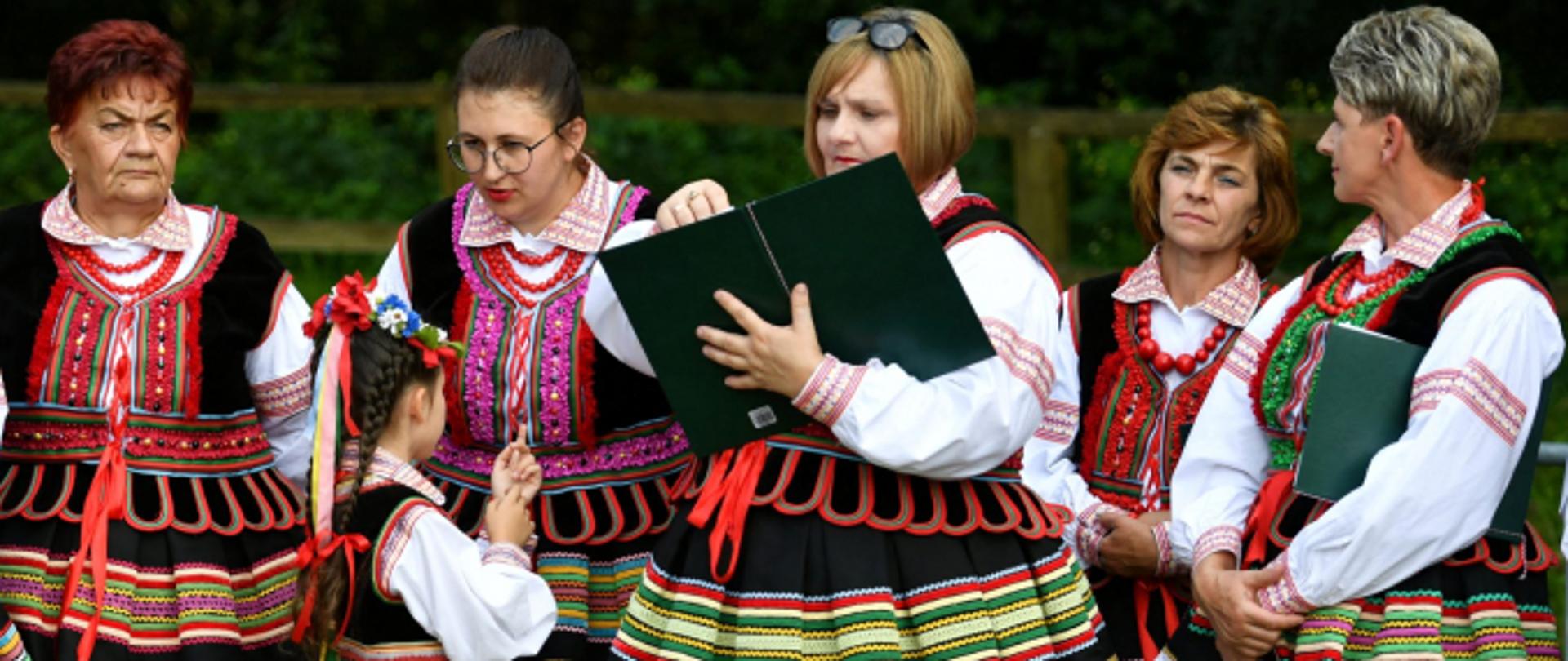 Na fotografii 5 pań ubranych w kolorowe stroje ludowe. Koło jednej z pań stoi dziewczynka ubrana również w strój ludowy. Jedna z kobiet trzyma zieloną okładkę. 