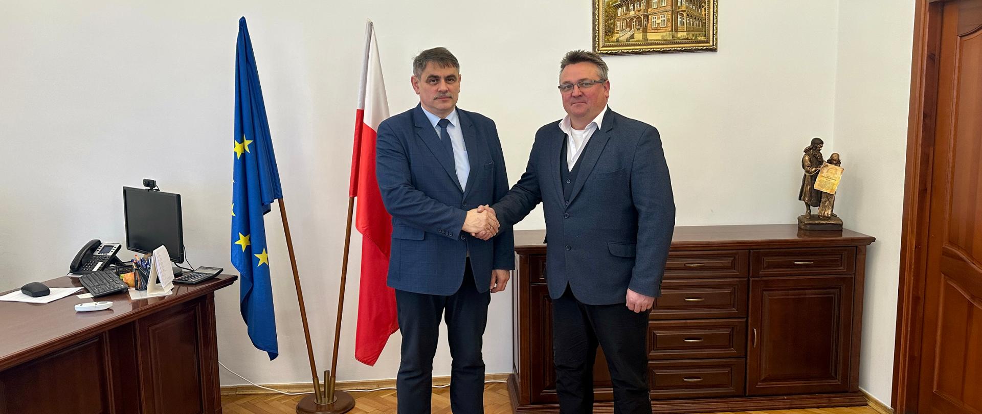 Dwóch mężczyzn w garniturach podaje sobie dłonie na tle polskiej i unijnej flagi w urzędowym gabinecie. Z lewej strony stoi biurko z komputerem i telefonem, z prawej komoda z figurką. Nad nimi na ścianie widoczny jest godło Polski oraz obraz przedstawiający budynek w stylu drewnianym.