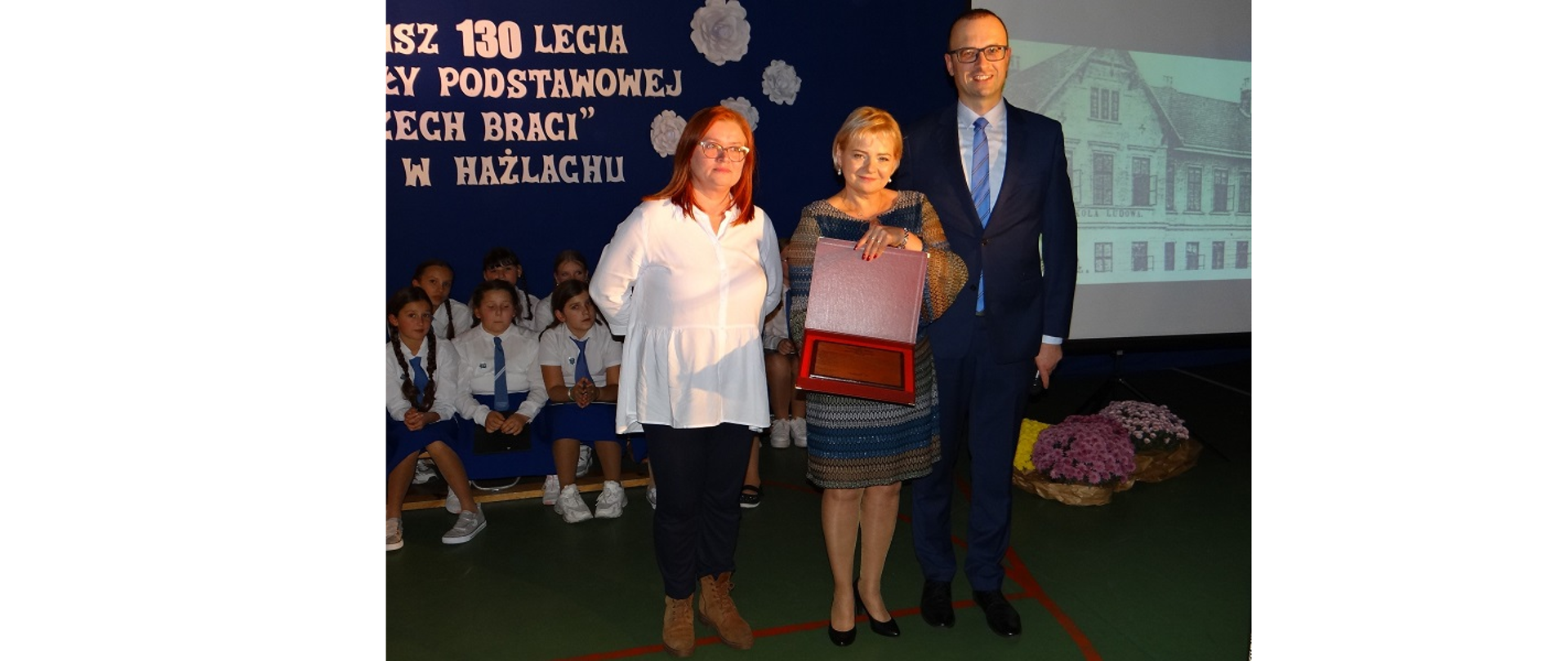 Dyrektor Aneta Milanowska-Trojszczak prezentuje pamiątkowy dyplom z życzeniami od władz Gminy Hażlach. Na zdjęciu stoją trzy osoby. Dyplom w ozdobnym etui prezentuje kobieta stojąca w środku