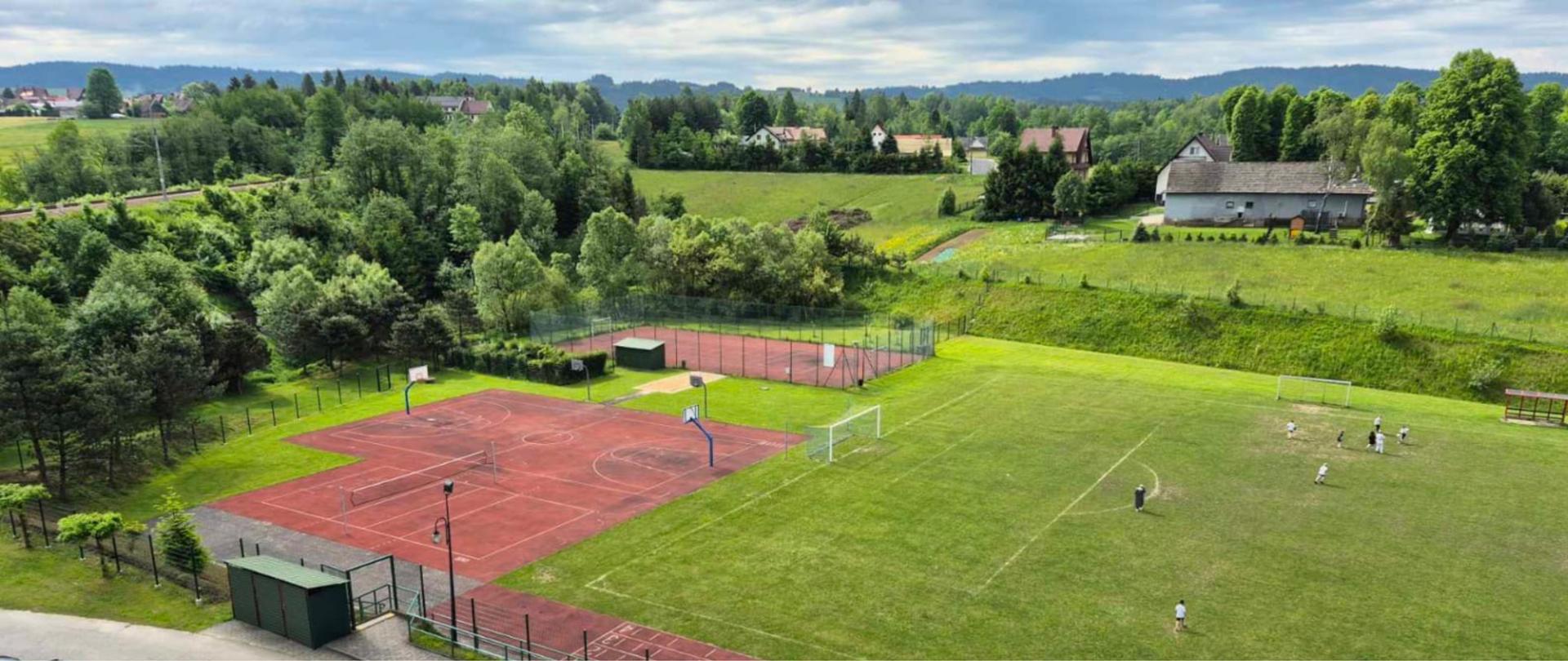 Widok z lotu ptaka na kompleks sportowo-rekreacyjny na terenach wiejskich. Na pierwszym planie znajdują się czerwone boiska wielofunkcyjne do koszykówki i tenisa oraz duże trawiaste boisko piłkarskie, na którym kilka osób ćwiczy lub gra. Obiekt otaczają drzewa, łąki i pola, a w tle widoczne są rozproszone zabudowania oraz zalesione wzgórza pod pochmurnym niebem.