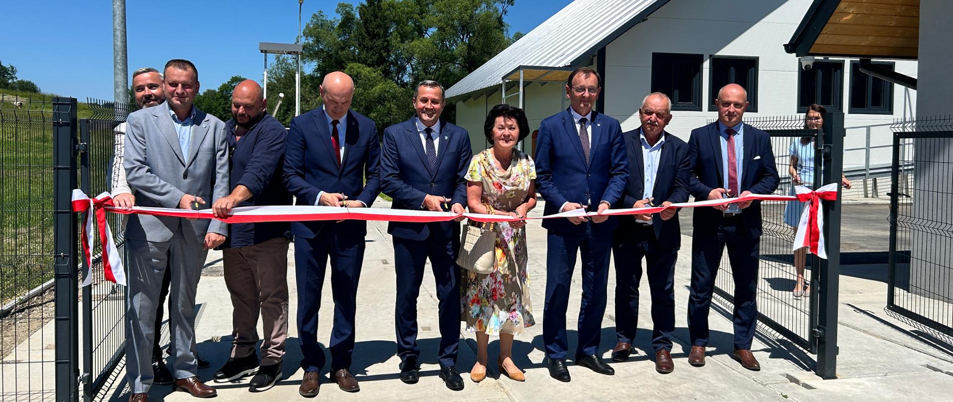 Grupa osób przecina wstążkę biało czerwoną zawieszoną na bramie.