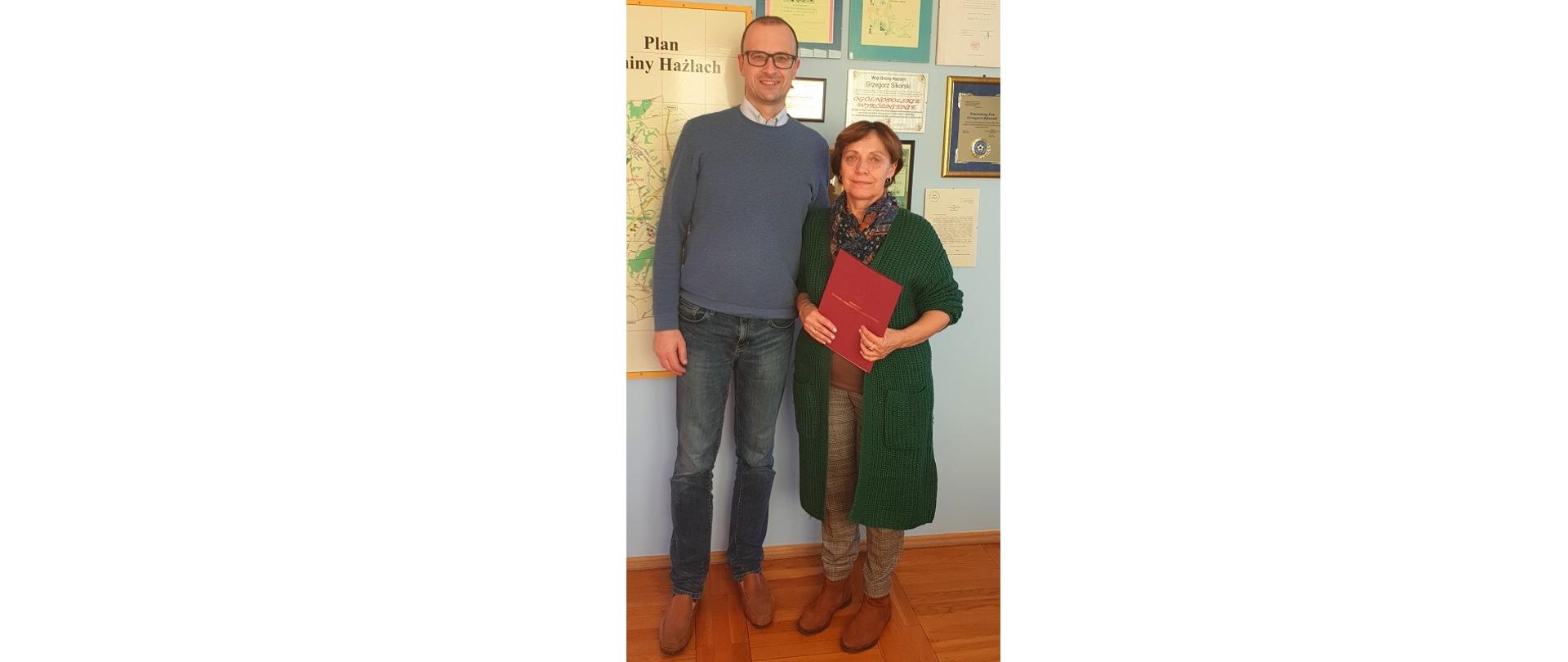Krystyna Penkała z Wójtem Gminy Hażlach Grzegorzem Sikorskim. Zdjęcie wykonane w gabinecie wójta w Urzędzie Gminy Hażlach. Osoby pozują do fotografii na stojąco. Kobieta trzyma w ręku ozdobną teczkę z napisem: Minister Kultury i Dziedzictwa Narodowego