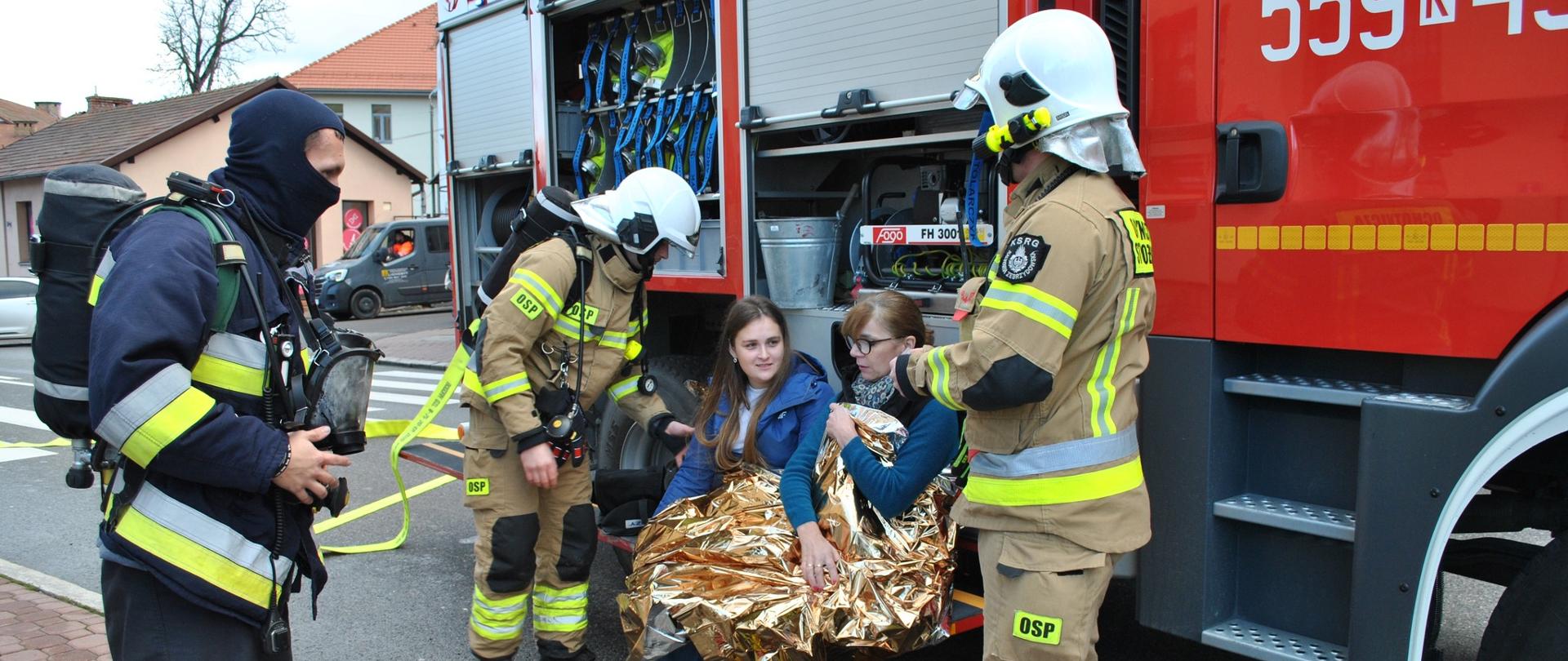 Na zdjęciu kilku strażaków w pełnym umundurowaniu ratowniczym udziela pomocy dwóm kobietom siedzącym na stopniu wozu strażackiego. Kobiety są okryte folią termoizolacyjną i wyglądają na poszkodowane, ale przytomne. Strażacy sprawdzają ich stan i zabezpieczają miejsce zdarzenia. Na ziemi leży otwarta torba medyczna z wyposażeniem, a w tle widoczny jest czerwony samochód ratowniczo-gaśniczy z uchylonymi schowkami sprzętowymi. Scena wygląda na ćwiczenia lub działania ratownicze w przestrzeni miejskiej.