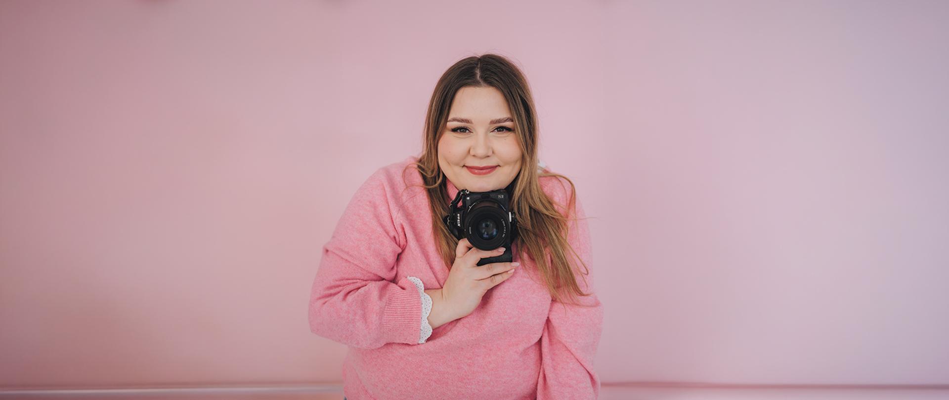 Uśmiechnięta kobieta w różowym swetrze trzyma aparat fotograficzny. Zdjęcie na różowym tle.