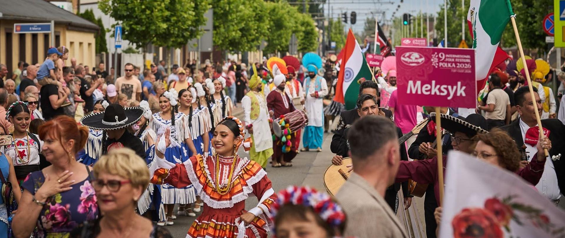 Uliczna parada podczas festiwalu Polka w Konstantynowie Łódzkim. Kolorowy korowód tancerzy w strojach ludowych z Meksyku i Indii maszeruje ulicą miasta. Widoczne flagi narodowe, tabliczki z nazwami państw oraz liczna publiczność. W tle widoczna miejska zabudowa.