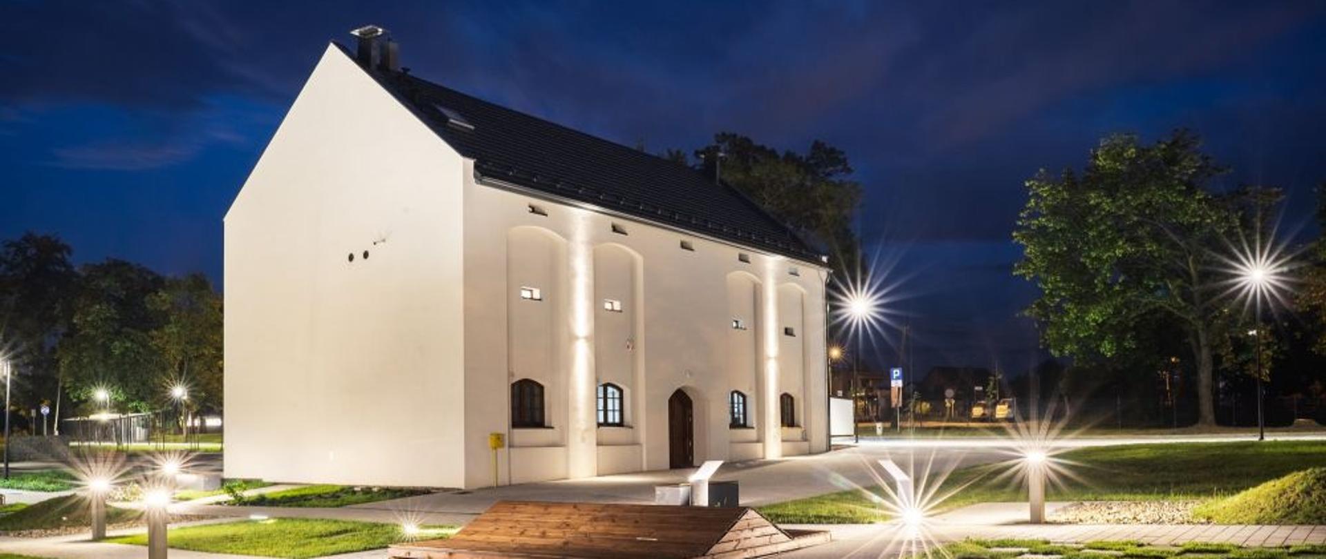 W środku zdjęcia odrestaurowany budynek byłego spichlerza nazwany Domem Przyrodnika. Wokół plac z zielenią. Fotografię wykonano w porze wieczornej, całość oświetlona sztucznym oświetleniem nocnym. 