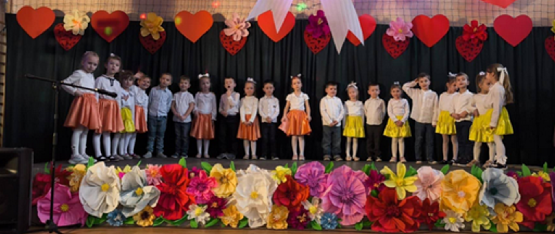 Grupa kilkunastu dzieci stoi na scenie podczas uroczystości szkolnej z okazji Dnia Babci i Dziadka. Dzieci ubrane są odświętnie – dziewczynki w kolorowych spódniczkach, chłopcy w jasnych koszulach. Scena udekorowana jest motywami serc, kwiatów oraz dużą dekoracją w kształcie serca z białą kokardą zawieszoną centralnie nad występującymi. Na pierwszym planie widoczne są barwne, papierowe kwiaty zdobiące front sceny.