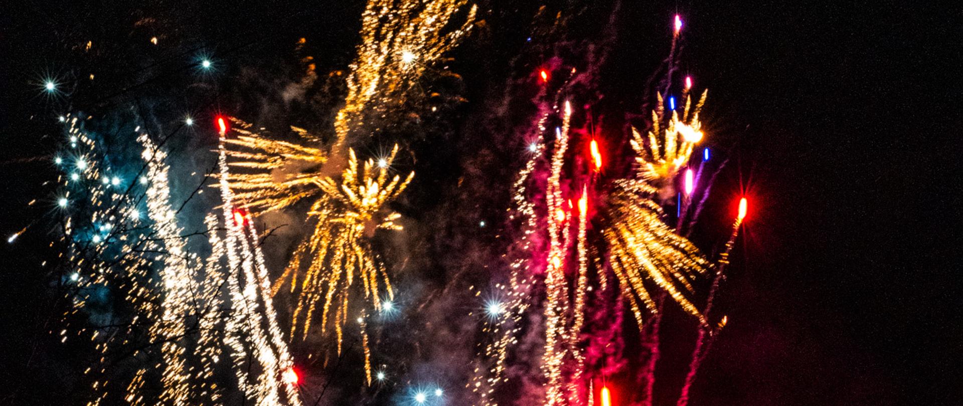 Pokaz bezhukowych sztucznych ogni podczas sylwestrowego powitania Nowego Roku na Rynku w Kalwarii Zebrzydowskiej. Kolorowe fajerwerki rozświetlają nocne niebo, a zgromadzeni mieszkańcy obserwują widowisko i dokumentują je telefonami.