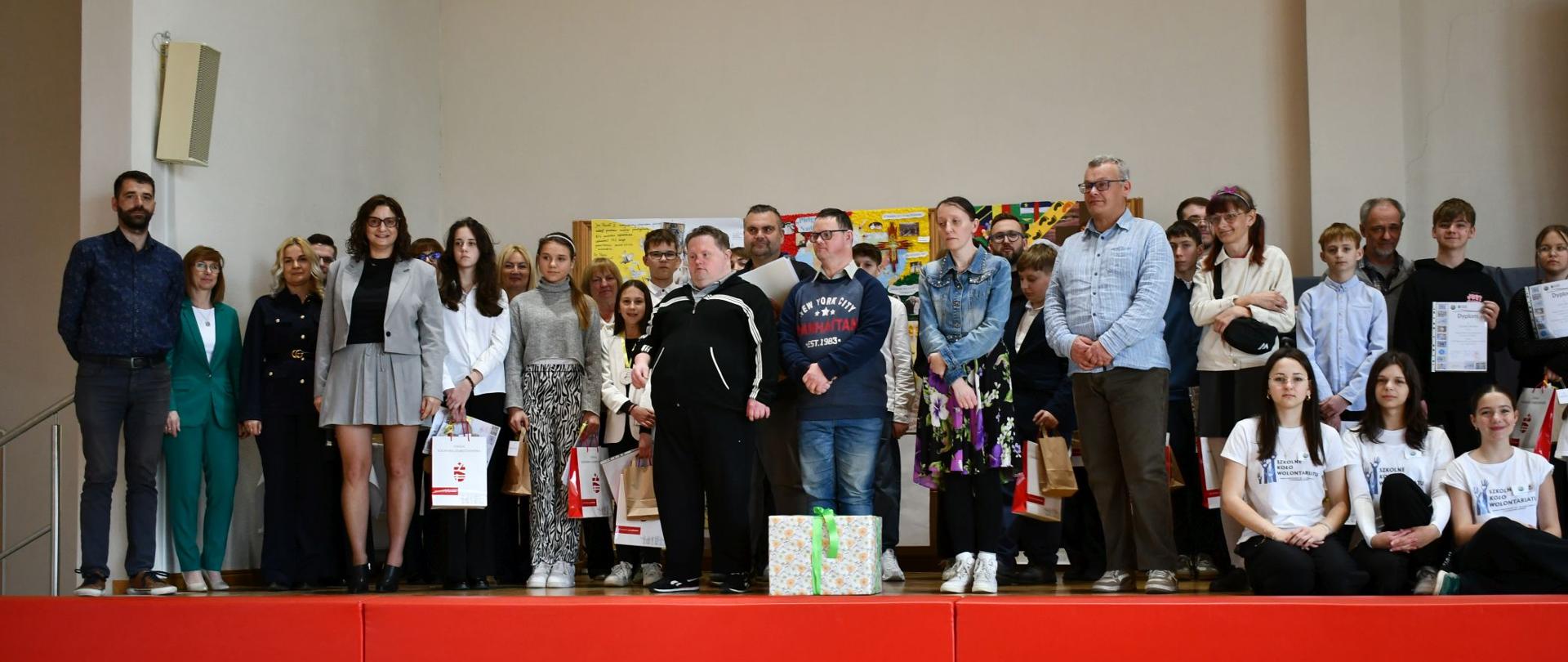 Grupowe zdjęcie wykonane na scenie podczas szkolnego wydarzenia. Na podwyższeniu stoi kilkadziesiąt osób – uczniowie, nauczyciele oraz organizatorzy – ustawieni w jednym rzędzie. Część uczestników trzyma dyplomy i torby z nagrodami, co wskazuje na zakończenie konkursu lub uroczystości wręczenia wyróżnień. W tle widoczne są kolorowe plansze i prace uczniów. Na pierwszym planie znajduje się czerwone podium, a po prawej stronie kilka osób siedzi, ubranych w jednakowe koszulki. Atmosfera jest oficjalna, ale jednocześnie swobodna i radosna, charakterystyczna dla szkolnych podsumowań i wspólnych zdjęć.