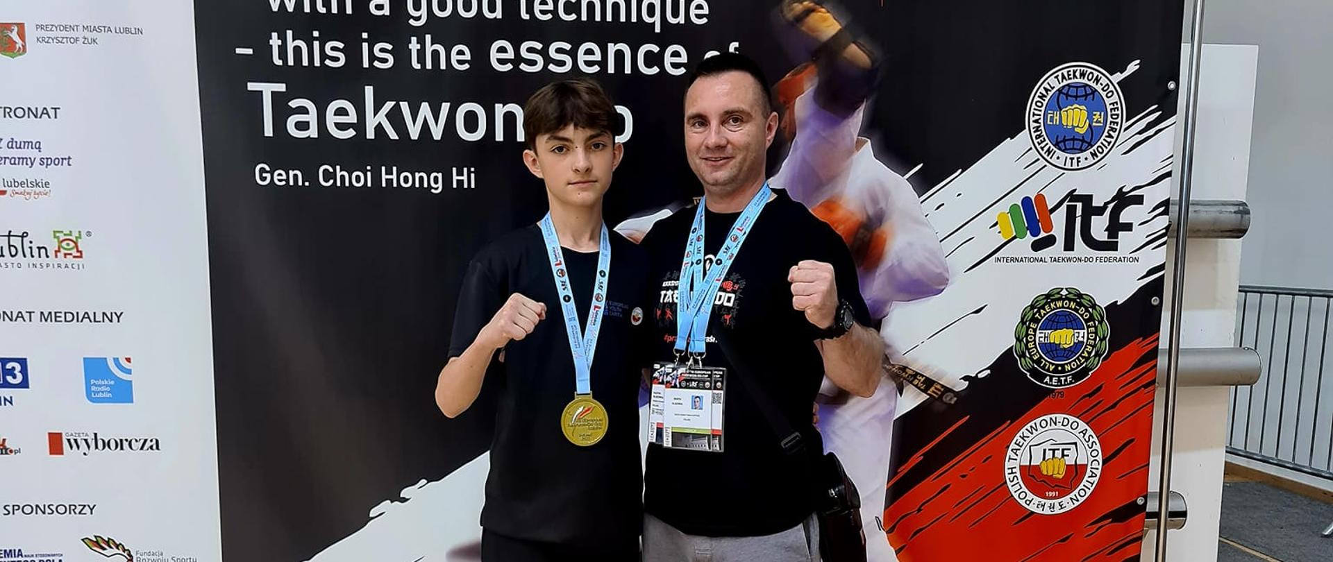 Kacper Olszówka wraz z medalem z zawodów zawieszonym na szyi. Obok niego od prawej strony stoi jego tata i jednocześnie trener Kraśnickiej Akademii Taekwon-do Marcin Olszówka