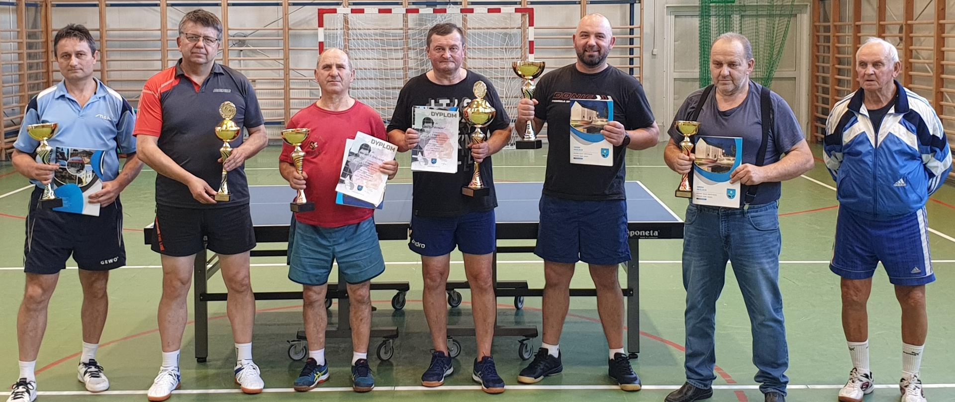 Zwycięzcy memoriału wraz ze współorganizatorem turnieju Józefem Michalikiem (pierwszy z prawej). Na fotografii 6 mężczyzn w różnym wieku prezentuje puchary i dyplomy. Z tyłu za nimi stół do tenisa stołowego. 