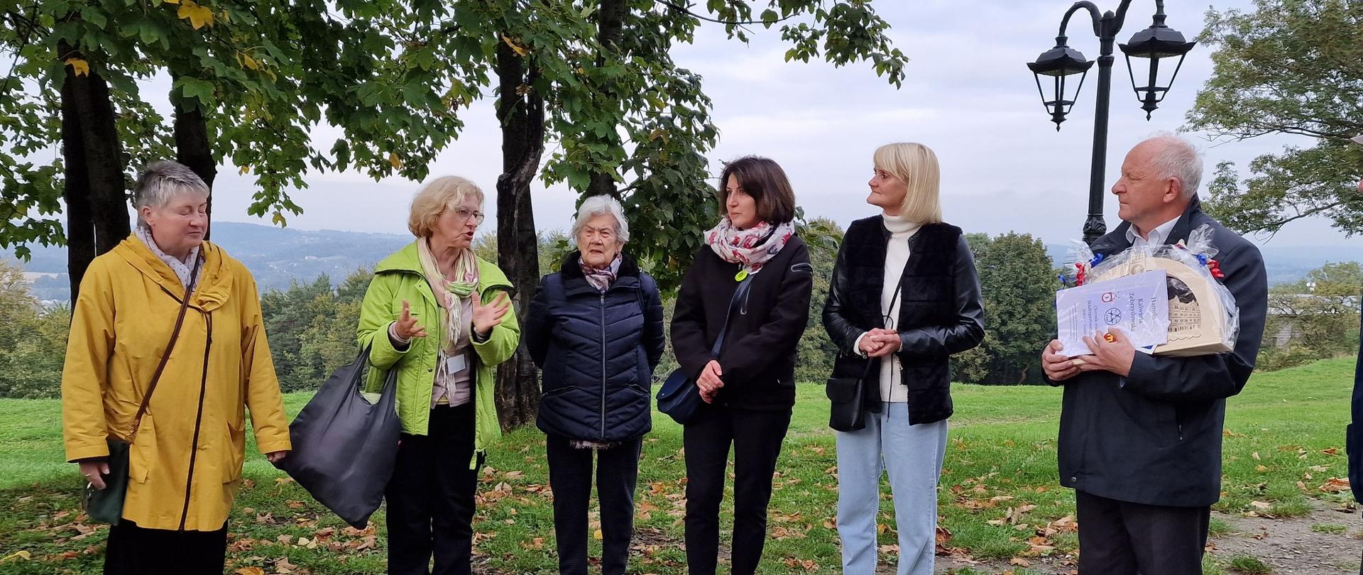 Fotografia w plenerze – częściowo na trawie, częściowo na ubitej ziemi w otoczeniu drzew stoją w rzędzie ciepło ubrani uczestnicy spotkania i rozmawiają. Burmistrz trzyma podarunki dla gości.