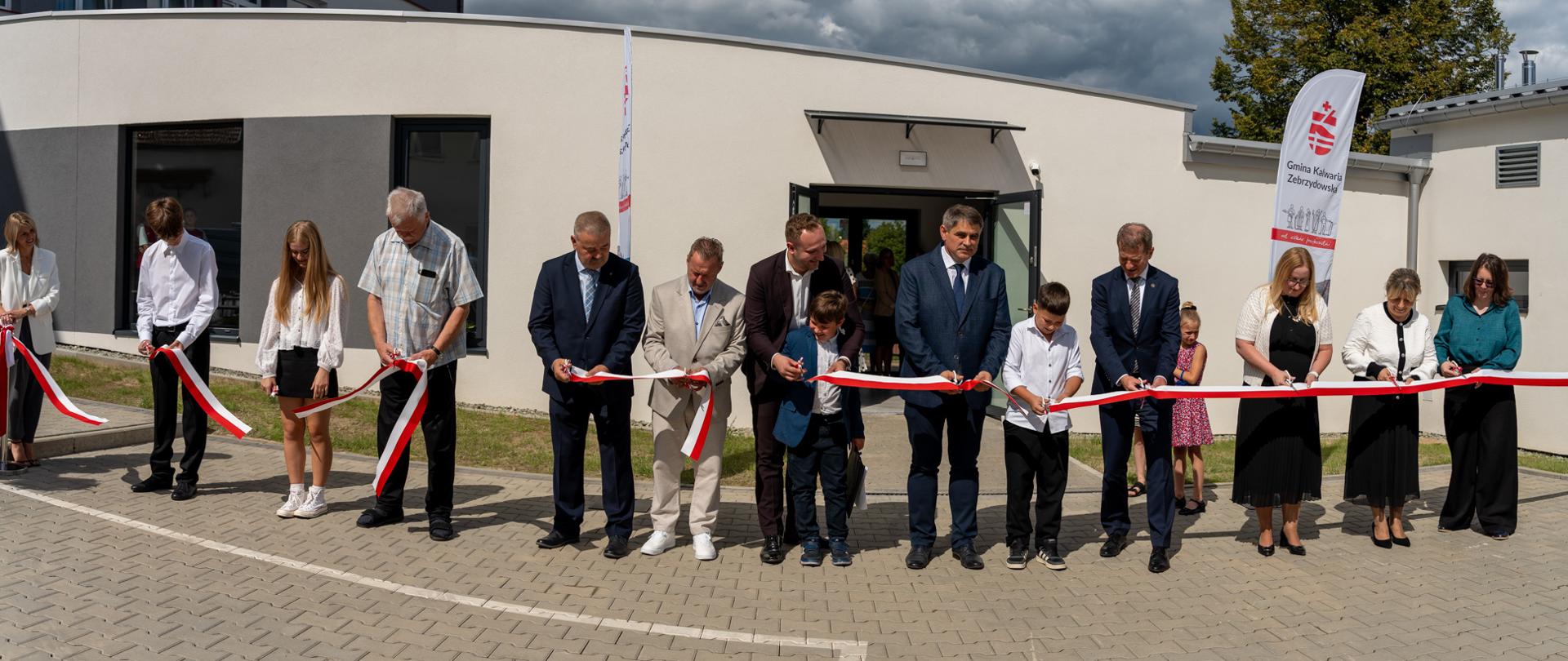 Uroczyste przecięcie biało-czerwonej wstęgi podczas otwarcia nowej sali gimnastycznej przy Zespole Szkół nr 7 w Barwałdzie Średnim. W ceremonii przed budynkiem uczestniczą przedstawiciele władz, zaproszeni goście, pracownicy szkoły oraz uczniowie. W tle widoczna jest flaga Gminy Kalwaria Zebrzydowska. Uroczystość odbyła się 1 września 2026 roku. Fot. Andrzej Famielec.