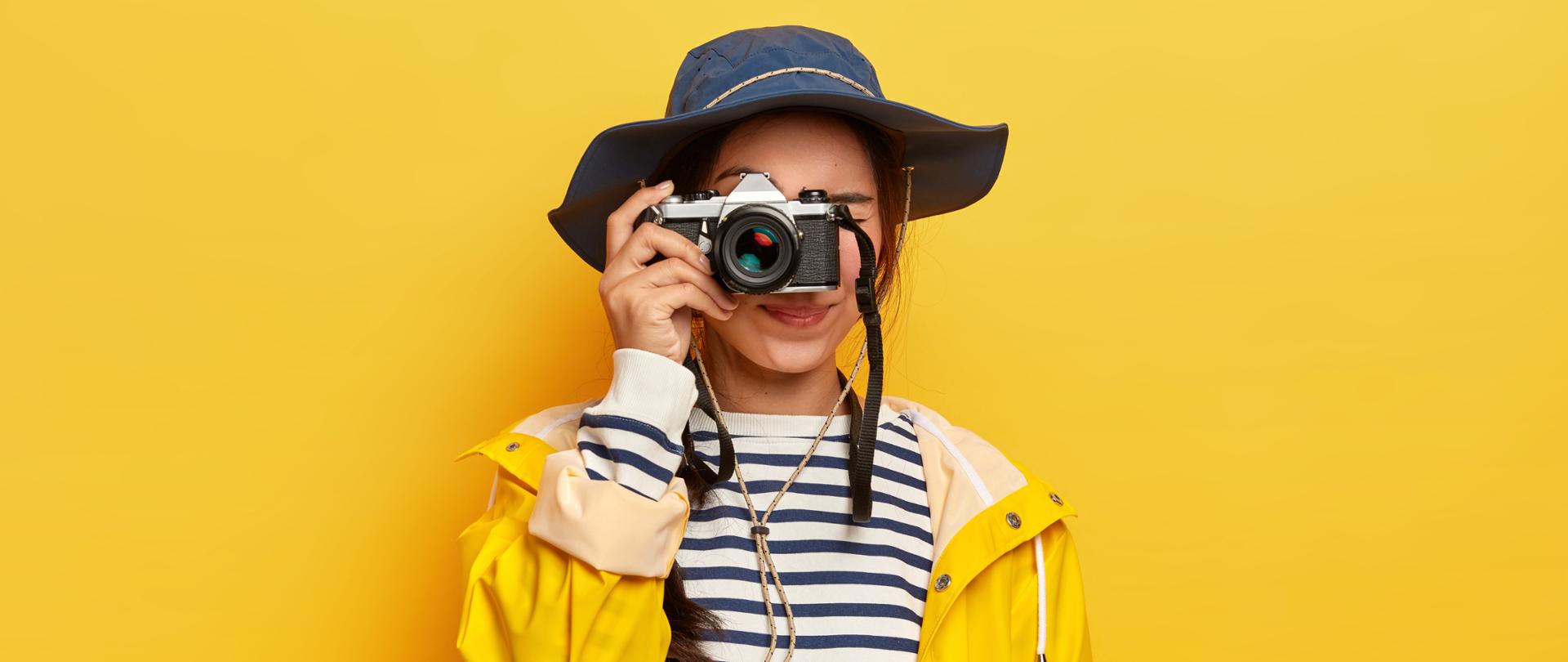 Na żółtym tle stoi dziewczynka w żółtym płaszczu. Ma bluzkę w paski, brązowe włosy, granatowy kapelusz i aparat fotograficzny w ręku.