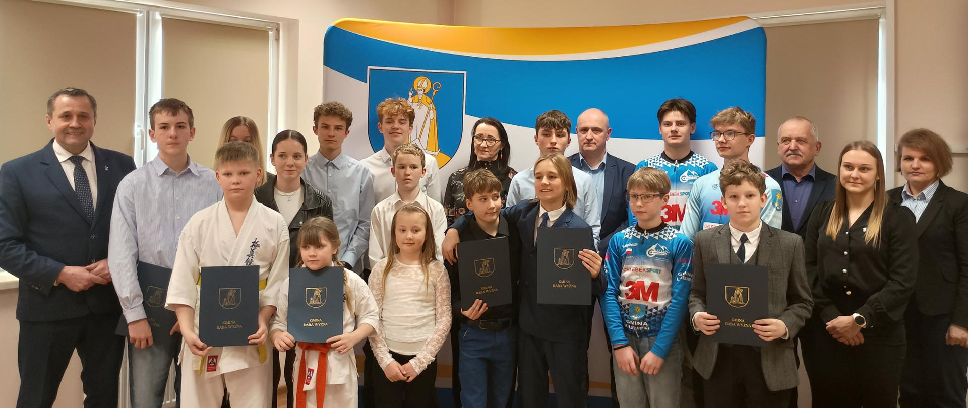 Grupa sportowców z dyplomami wraz z Panem Wójtem Gminy Raba Wyżna, Panią Skarbik, Panem Przewodniczącym Rady Gminy i członkowie Komisji Oświaty, Kultury, Sportu i Ochrony Socjalnej Rady Gminy Raba Wyżna stoją na tle ścianki Gminy Raba Wyżna 