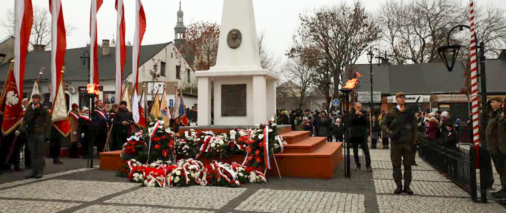 Na zdjęciu biały pomnik poświęcony Marszałkowi Józefowi Piłsudskiemu znajdujący się w Kraśniku. Pod pomnikiem zostały złożone wieńce z biało-czerwonymi kwiatami i wstęgami. Przy pomniku stoją żołnierze pełniący honorową wartę. Na lewo od pomnika stoją poczty sztandarowe a za nimi zawieszone na masztach biało-czerwone flagi. Na prawo i za pomnikiem zgromadzeni są uczestnicy wydarzenia.