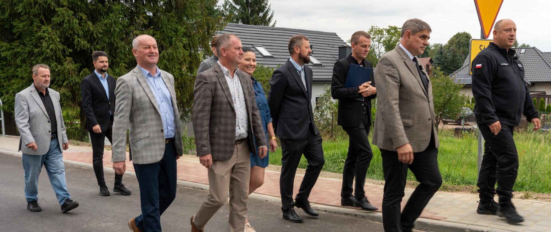 Grupa przedstawicieli administracji rządowej i samorządowej podczas wizyty wojewody małopolskiego Krzysztofa Jana Klęczara w Przytkowicach. Uczestnicy spotkania idą wspólnie przebudowaną drogą gminną.