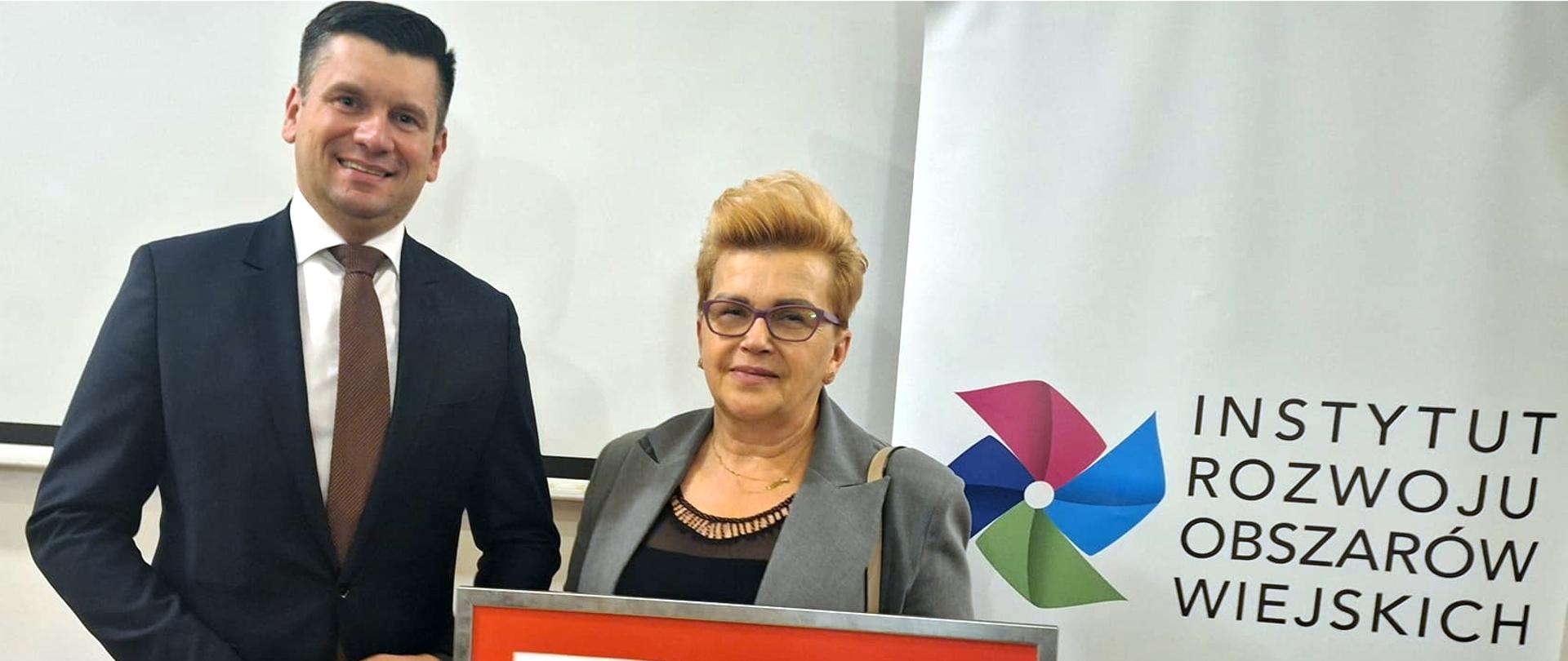 Sołtys Ewa Pacut stoi koło Wicemarszałka Łukasza Smółki trzymając oprawiony w ramę czek z napisem Małopolska Wieś 2023, w tle baner z logo instytutu rozwoju obszarów wiejskich oraz takim też napisem