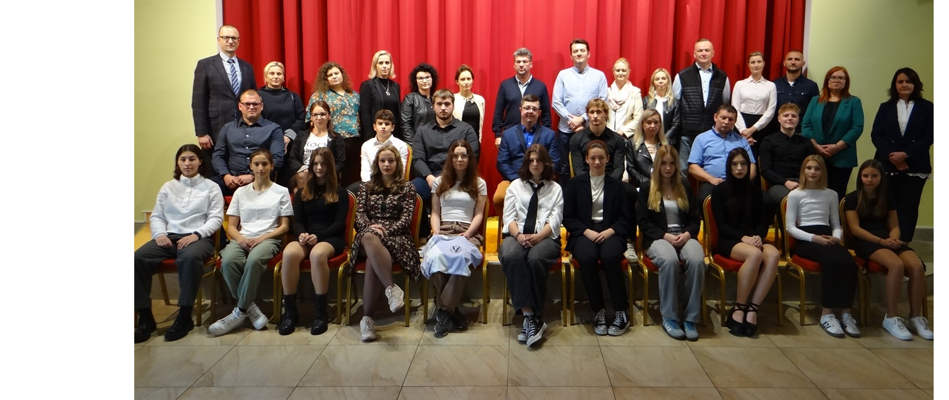 Pamiątkowa fotografia młodych sportowców z rodzicami i przedstawicielami Gminy Hażlach. Na zdjęciu wykonanym w sali widowiskowej Gminnej Biblioteki Publicznej w Hażlachu brakuje m.in. zawodników UKS Nakano Pogwizdów, którzy w tym dniu brali udział w zawodach. Osoby pozujące do fotografii siedzą w dwóch rzędach na krzesłach oraz stoją na scenie
