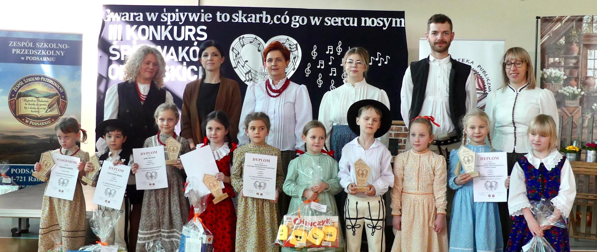 Grupa stojących dorosłych i dzieci, ubranych w stroje regionalne. Dzieci trzymają dyplomy oraz nagrody.