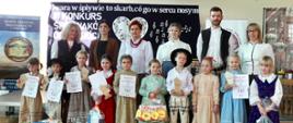 Grupa stojących dorosłych i dzieci, ubranych w stroje regionalne. Dzieci trzymają dyplomy oraz nagrody.