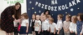 Przedszkolaki stoją w sali na tle ścianki "Pasowanie na przedszkolaka". Przed nimi stoi dziewczynka, recytuje do mikrofonu, który przytrzymuje prowadząca uroczystość