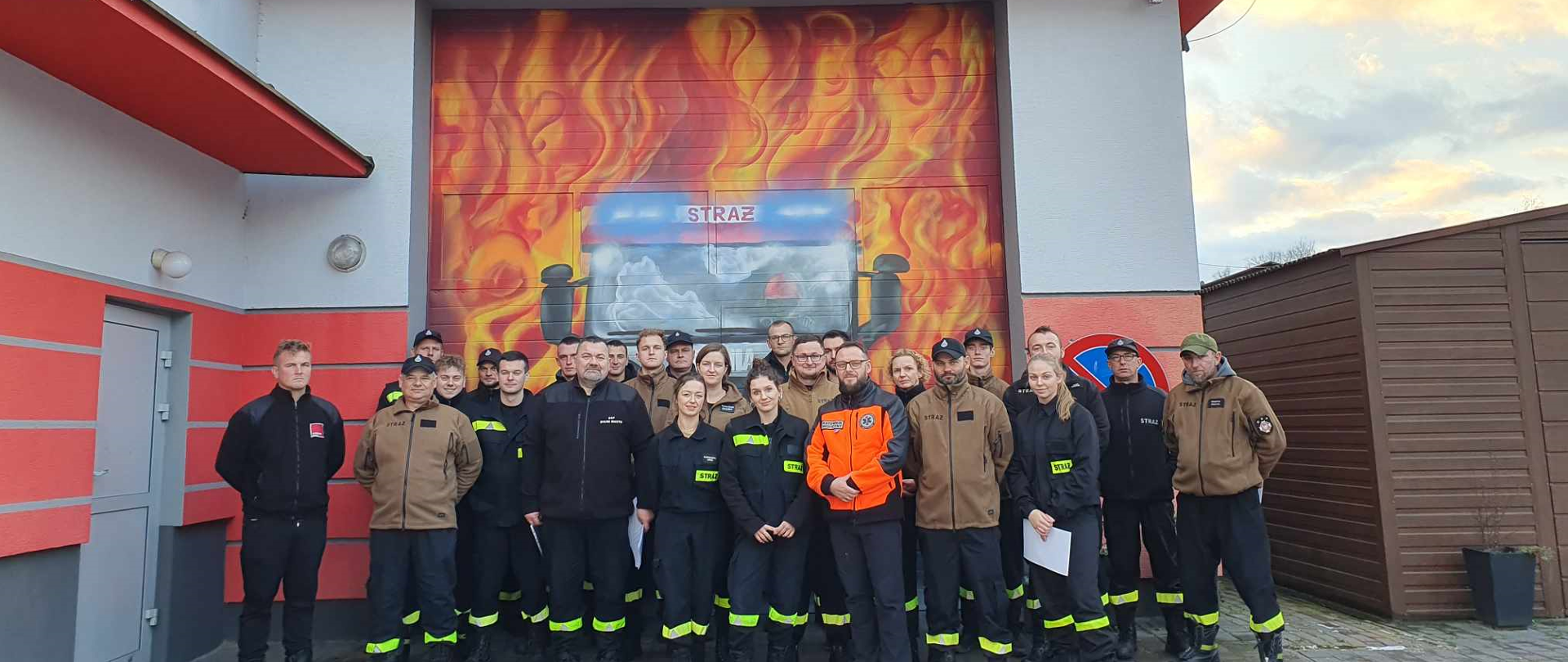 Na fotografii grupa strażaków ubranych w czarne oraz brązowo czarne stroje. Na drugim planie brama garażowa OSP Giedlarowa, na której namalowany został wóz strażacki w płomieniach. Powyżej bramy czerwony napis na szarym budynku Ochotnicza Straż Pożarna Giedlarowa. Z prawej strony brązowy garaż z lewej szare drzwi wejściowe. 