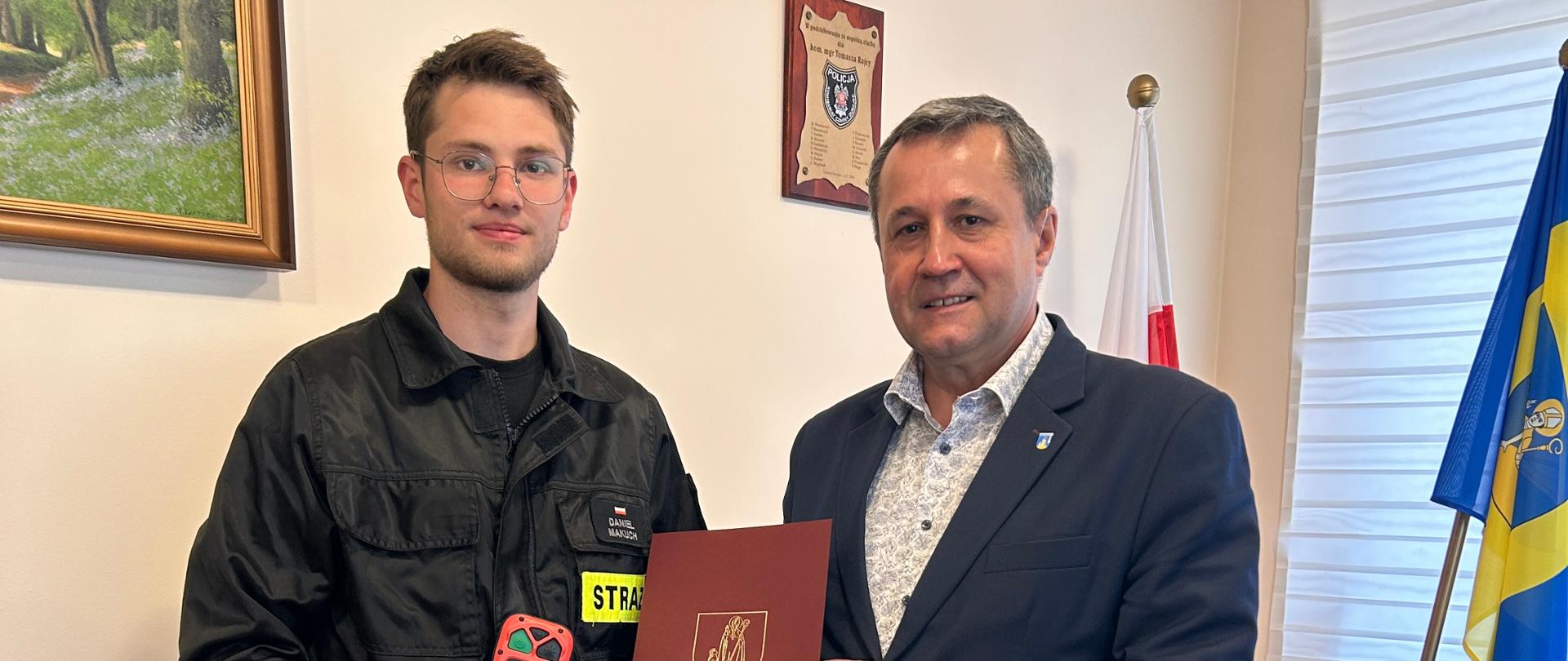 Strażak i przedstawiciel samorządu prezentują kamerę termowizyjną podczas oficjalnego przekazania sprzętu w pomieszczeniu biurowym. W tle flagi i jasna ściana z dwoma obrazami.