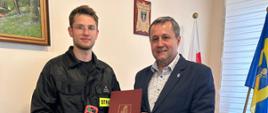 Strażak i przedstawiciel samorządu prezentują kamerę termowizyjną podczas oficjalnego przekazania sprzętu w pomieszczeniu biurowym. W tle flagi i jasna ściana z dwoma obrazami.