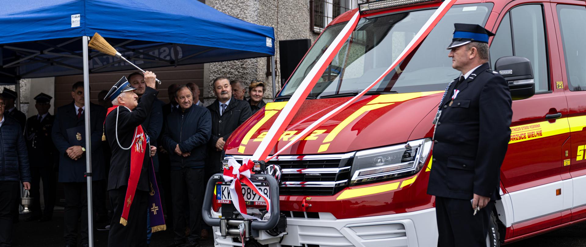 Na zdjęciu znajduje się ksiądz, który dokonuje poświecenia nowego samochodu dla OSP Zarzyce Wielkie. Na zdjęciu widoczni są również przedstawiciele lokalnych władz, strażacy oraz społecznicy. 
