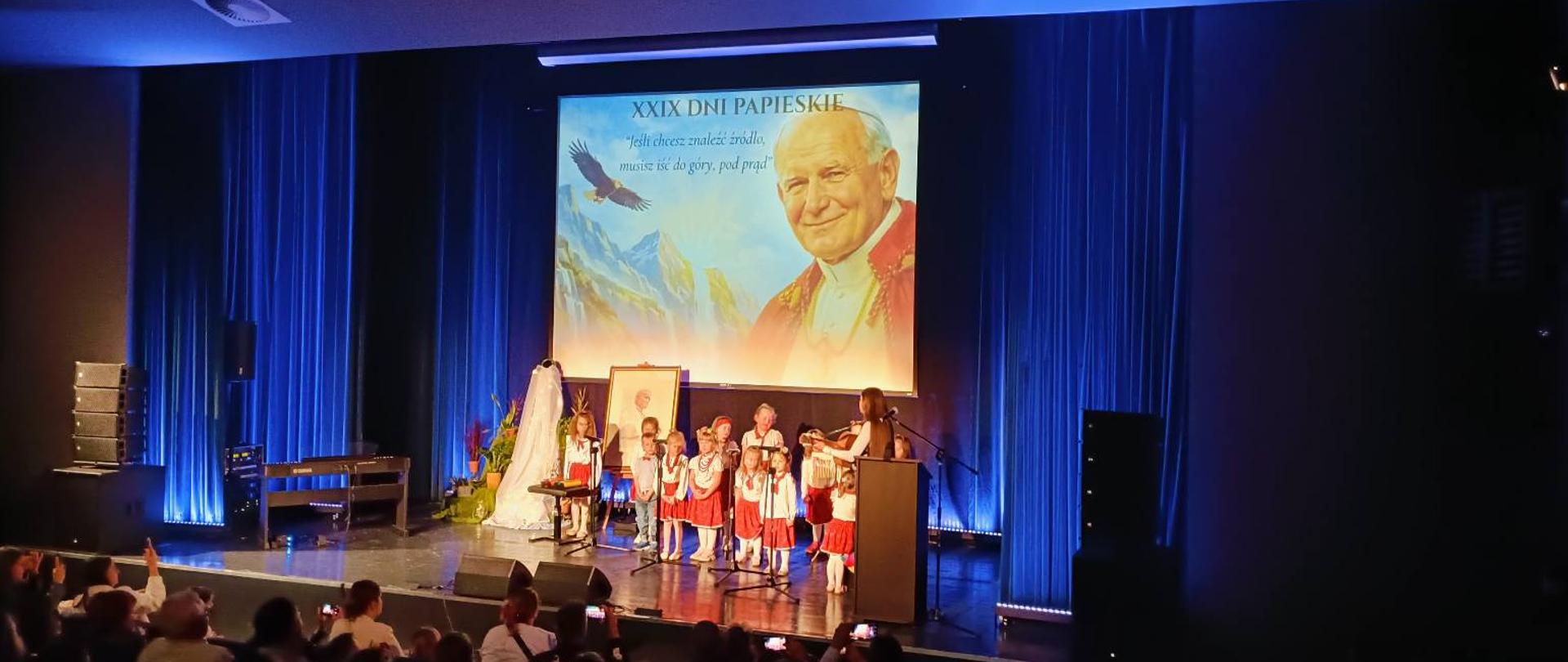 Na scenie sali widowiskowej odbywa się występ dzieci podczas Gali XXIX Dni Papieskich. Młodzi artyści ubrani w ludowe stroje stoją przed mikrofonami i prezentują program artystyczny. W tle widoczny jest duży ekran z wizerunkiem św. Jana Pawła II oraz hasłem przewodnim wydarzenia. Scena oświetlona jest niebieskim światłem, a na widowni zgromadzeni są uczestnicy i goście obserwujący występ.