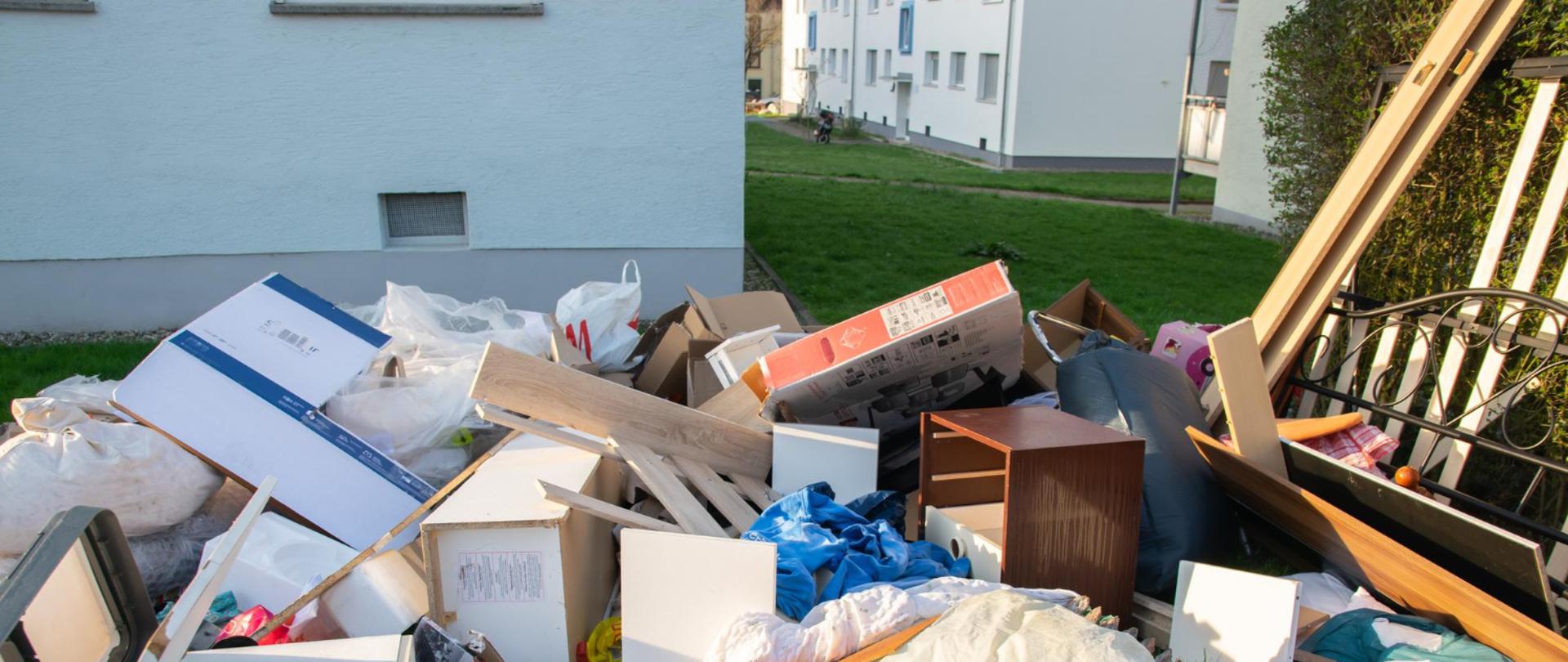Stos odpadów wielkogabarytowych przed budynkiem mieszkalnym. Widać stare meble, kartony, worki, pościel i elementy wyposażenia wnętrz.