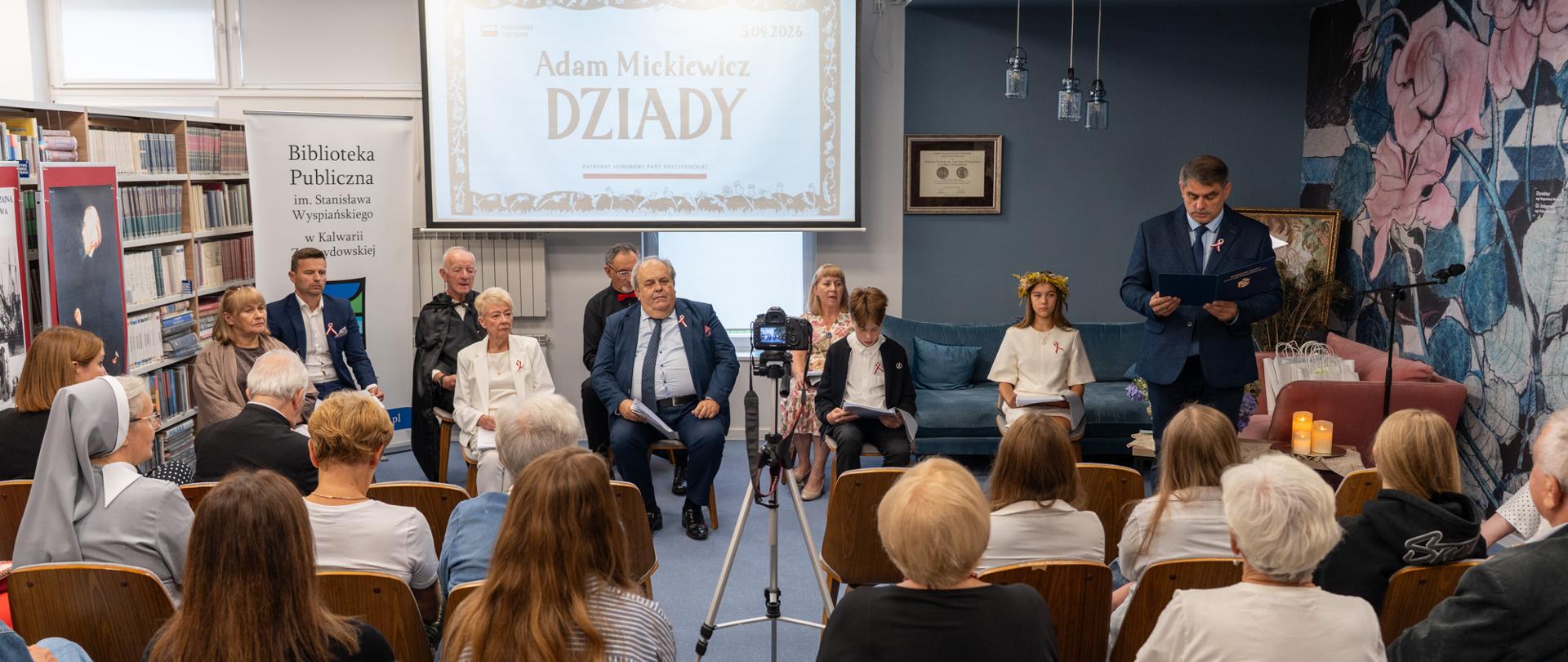 Uczestnicy Narodowego Czytania w Bibliotece Publicznej w Kalwarii Zebrzydowskiej. Na scenie zaproszeni goście odczytują fragmenty „Dziadów” Adama Mickiewicza, a wydarzenie obserwuje zgromadzona publiczność.