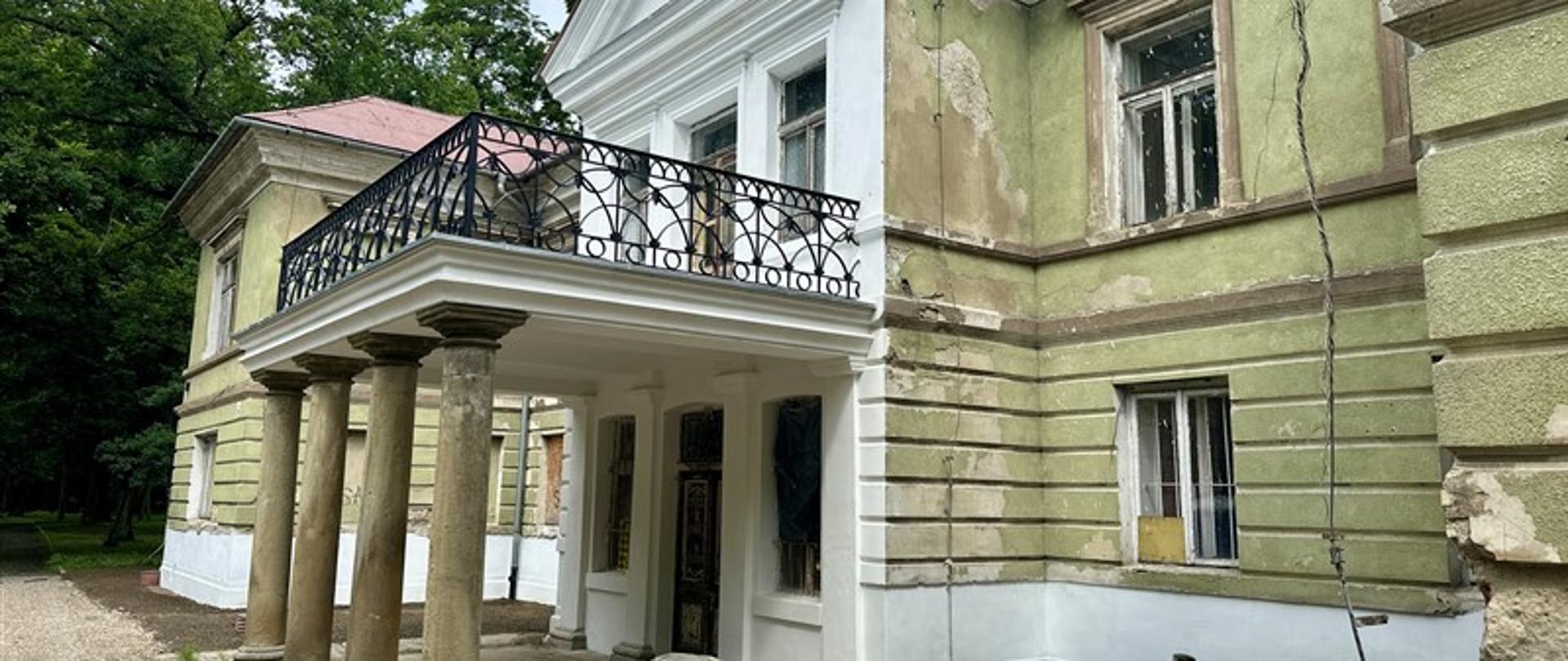 Zabytkowy dwór w trakcie prac renowacyjnych. Budynek o klasycystycznej formie z kolumnowym gankiem i balkonem z ozdobną, metalową balustradą. Elewacja częściowo odnowiona – fragmenty ścian są świeżo pomalowane na biało, inne nadal wymagają remontu, z widocznymi ubytkami tynku. Obiekt otoczony zielenią drzew, a przed wejściem znajduje się uporządkowany teren z ziemią i ścieżką żwirową.