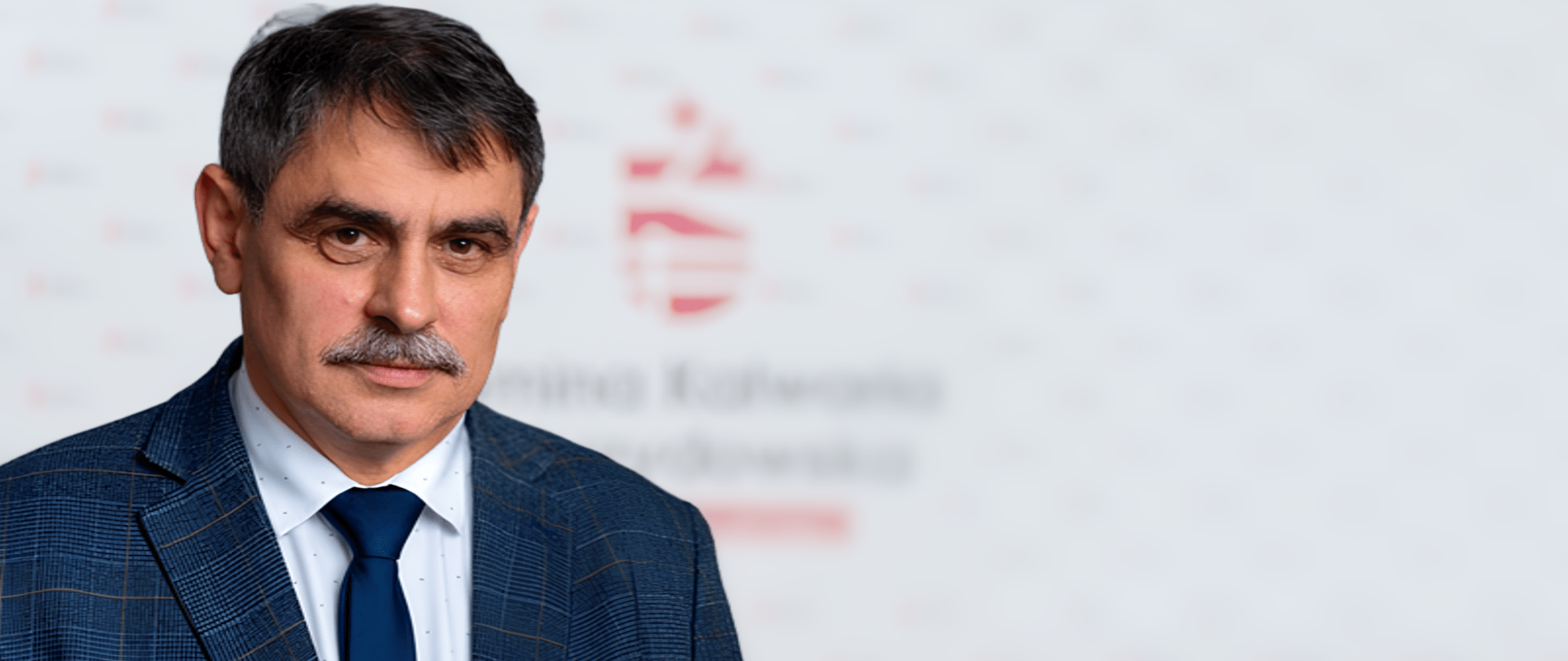 Fotografia portretowa – mężczyzna ubrany w garnitur w kratkę, białą koszulę i granatowy krawat, w tle logo Kalwarii Zebrzydowskiej