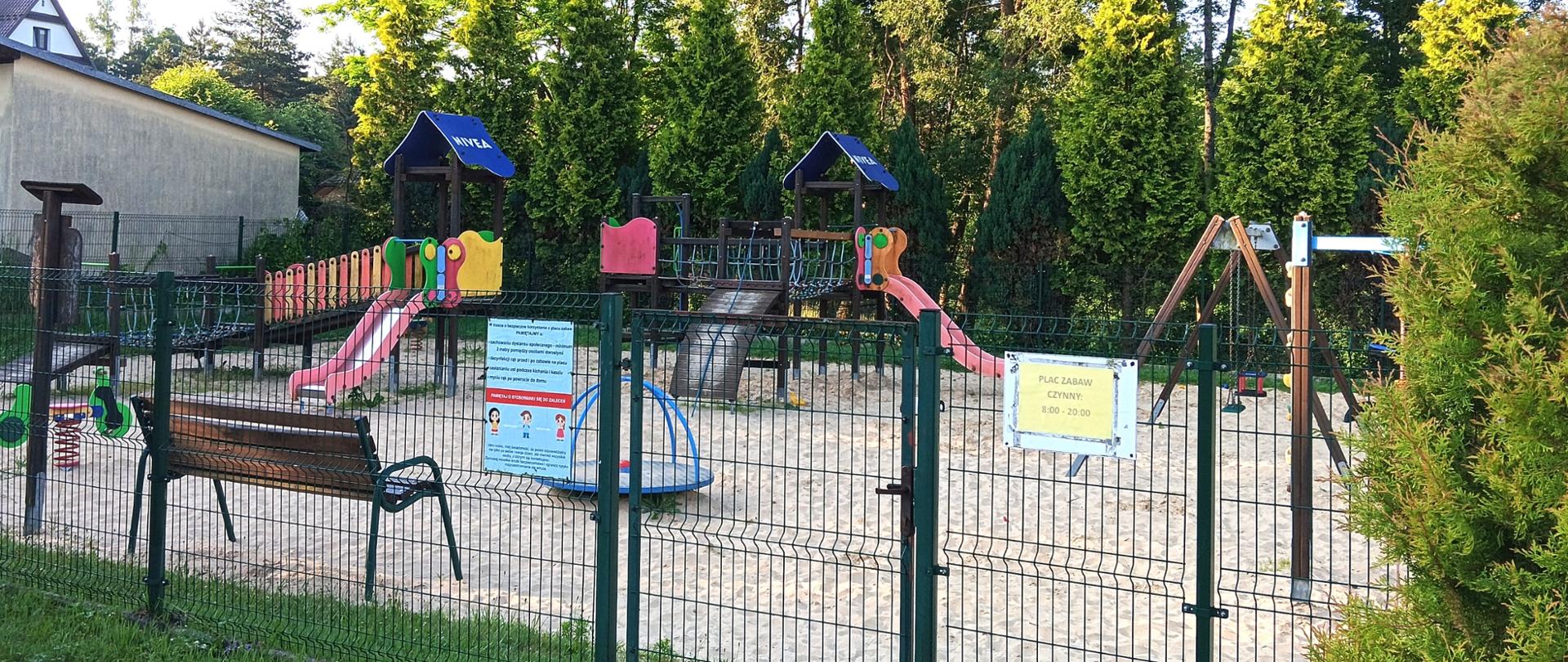 Widok na plac zabaw, ogrodzony z urządzeniami na nawierzchni z piasku, w tle drzewa.