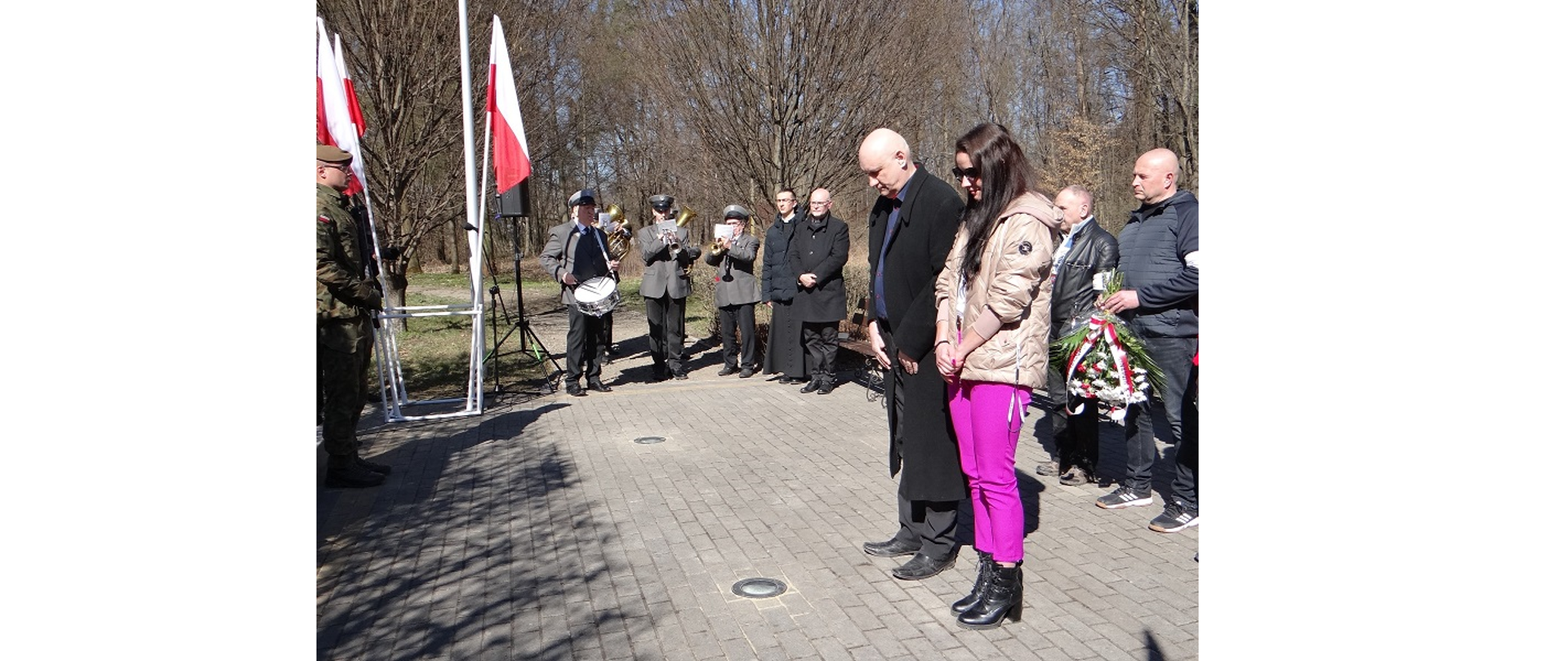 Przedstawiciele Gminy Hażlach oddają hołd pomordowanym żołnierzom ruchu oporu. Delegacje z kwiatami podchodzą kolejno przed pomnik, przed którym stoją żołnierze trzymający wartę honorową. W tle orkiestra i biało-czerwone flagi narodowe