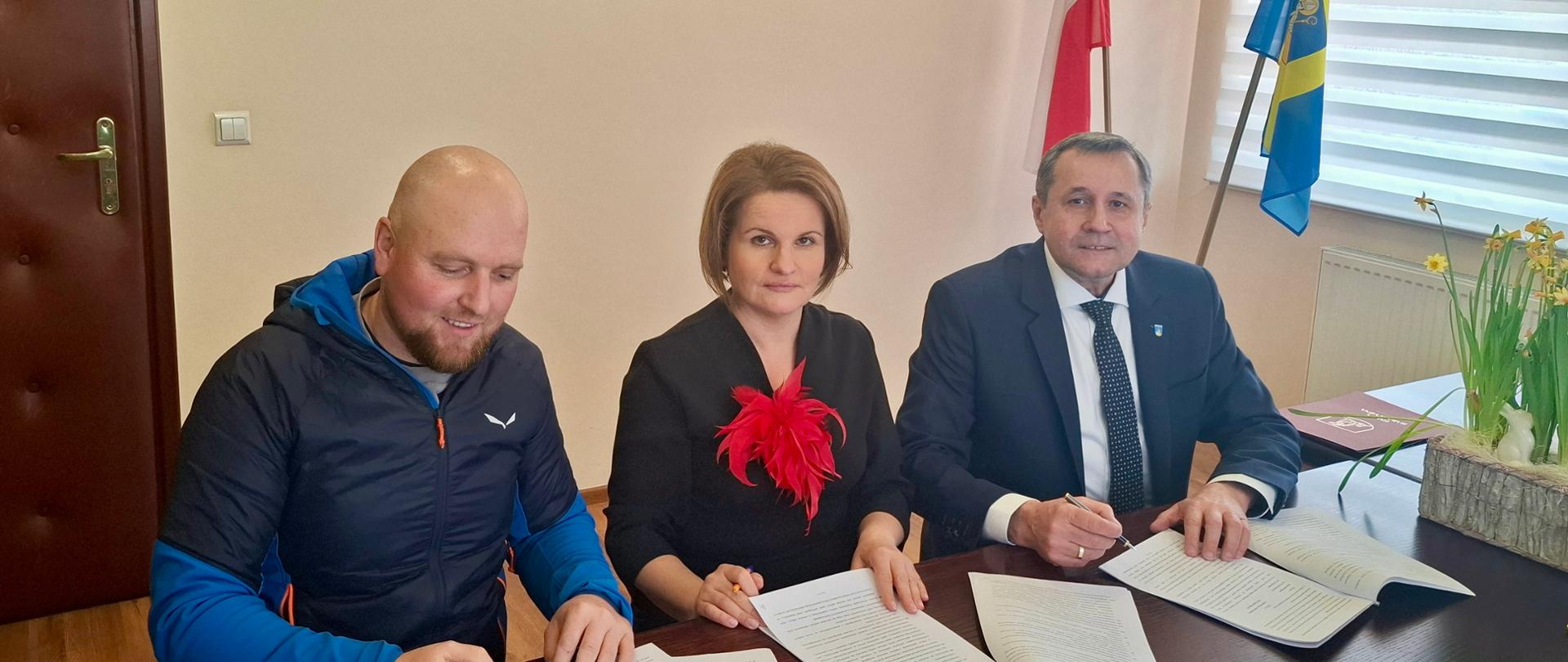 Na zdjęciu widoczne trzy osoby, siedzące za biurkiem z dokumentami. W tle ściana z obrazem i dwie flagi.