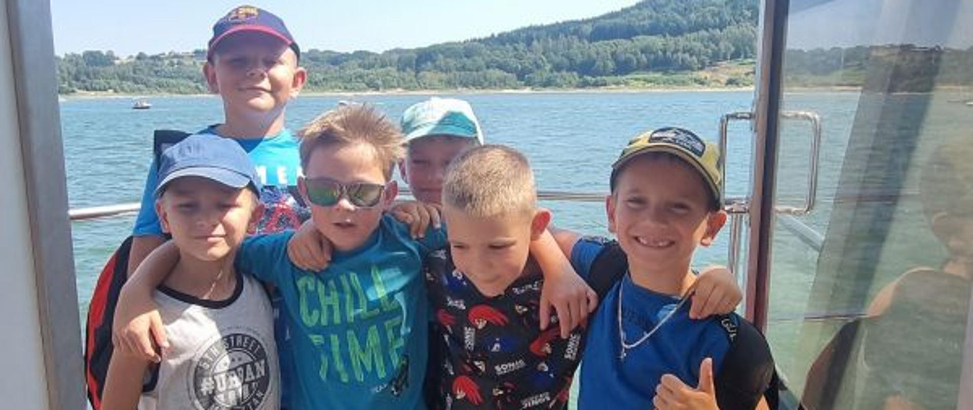 Grupa dzieci uczestniczących w półkolonii pozuje do wspólnego zdjęcia podczas rejsu statkiem. Uczestnicy stoją na pokładzie, obejmując się i uśmiechając do aparatu. W tle widoczne są jezioro, zielone wzgórza oraz bezchmurne, słoneczne niebo.