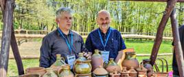 Na perwszym planie zdjęcia znajdują wyroby ceramiczne, za którymi stoją dwie osoby. Od lewej Cezary Gajewski oraz Andrzej Gotner. W tle za nimi znajduje się zagroda z drewna otoczona drzewami.