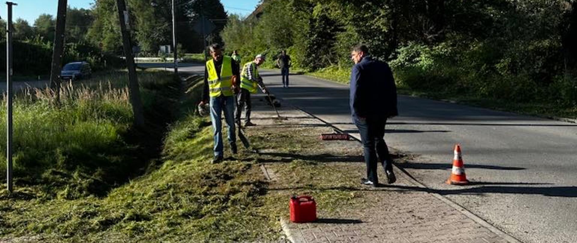 Na zdjęciu widać grupę osób pracujących przy poboczu drogi w słoneczny dzień. Kilku mężczyzn w odblaskowych kamizelkach wykonuje prace porządkowe, prawdopodobnie koszenie lub przycinanie trawy. Na chodniku i trawniku leżą resztki skoszonej roślinności, a w pobliżu stoi czerwony kanister. Obok pracowników znajduje się mężczyzna w ciemnym ubraniu, obserwujący działania. Na jezdni ustawiono pojedynczy pachołek ostrzegawczy. W tle widać linię energetyczną na drewnianym słupie, drzewa oraz fragment zabudowań, a niebo jest bezchmurne i intensywnie niebieskie.