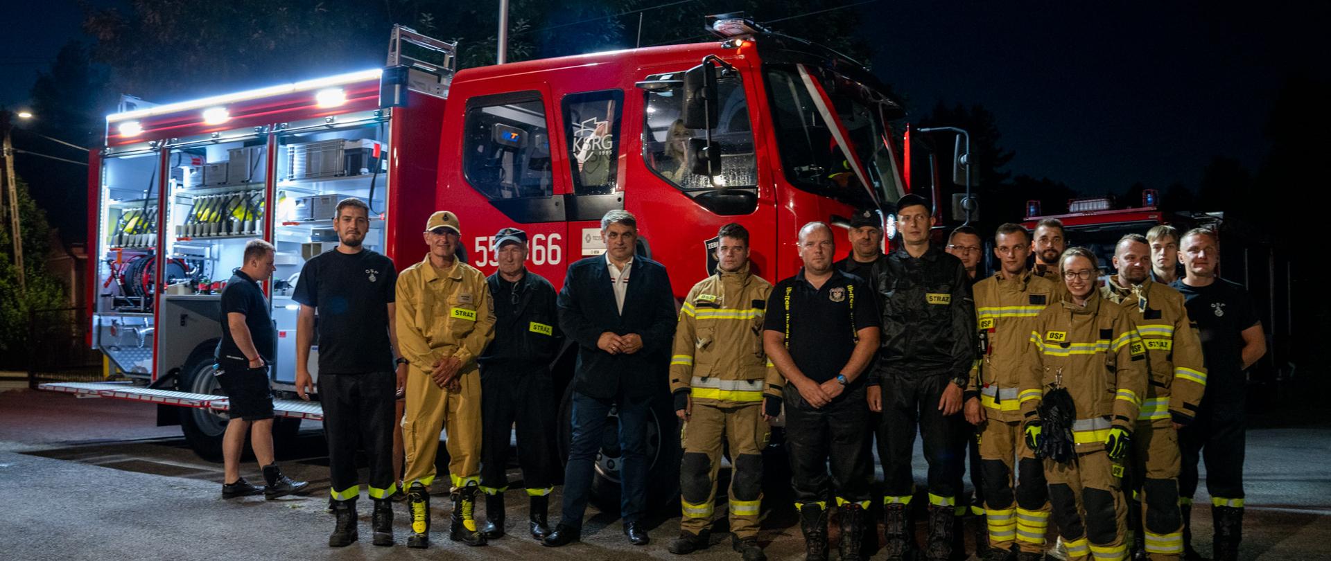 Na zdjęciu grupa strażaków pozuje do wspólnej fotografii przed nowoczesnym, czerwonym wozem strażackim, zaparkowanym na oświetlonym placu nocą. Część osób ubrana jest w pełne umundurowanie bojowe w kolorze beżowym lub czarnym z odblaskowymi pasami, inni w koszulki z emblematami straży. W tle, przy otwartych drzwiach pojazdu, widać wyposażenie ratownicze. Całą scenę oświetlają latarnie uliczne, a za strażakami widać zarys drzew i drugi wóz strażacki. 